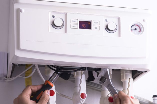 Person using a screwdriver and wrench to service a white boiler with gauges and control panel.
