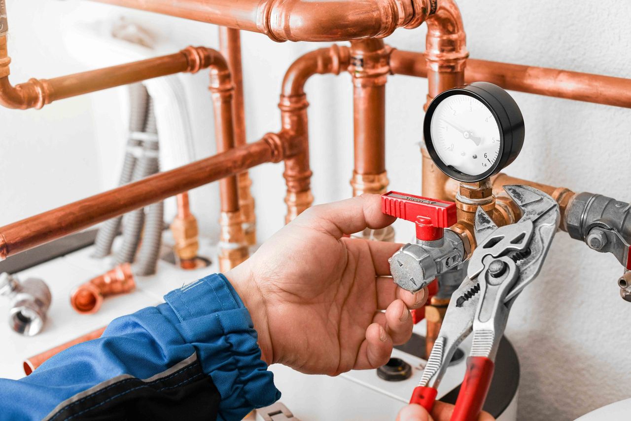 Plumber using pliers on valve with pressure gauge, copper pipes, in white setting.