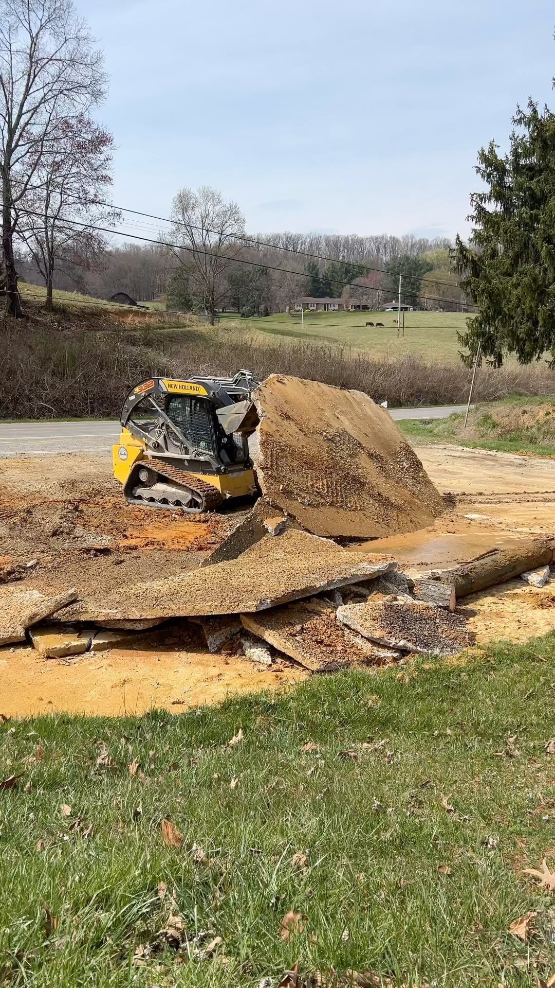 A yellow skid steer tractor demolishing asphalt in a grassy field on a sunny day.