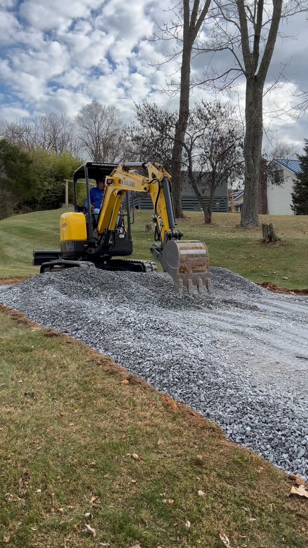 Yellow excavator spreading gravel on a driveway. Trees and house in the background.