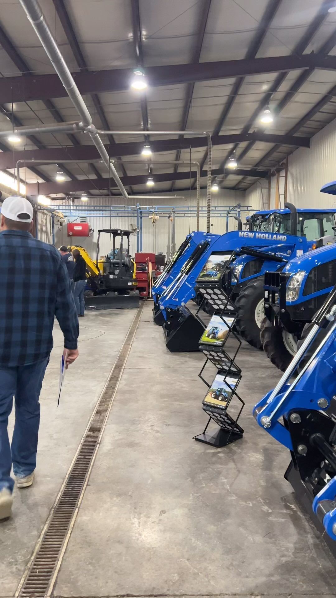 Person walks past blue tractors inside a warehouse. Display rack holds brochures.