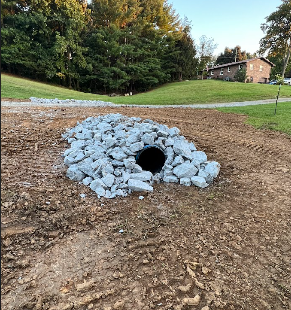 A culvert covered with rocks, in a dirt field with a house and trees in the background.