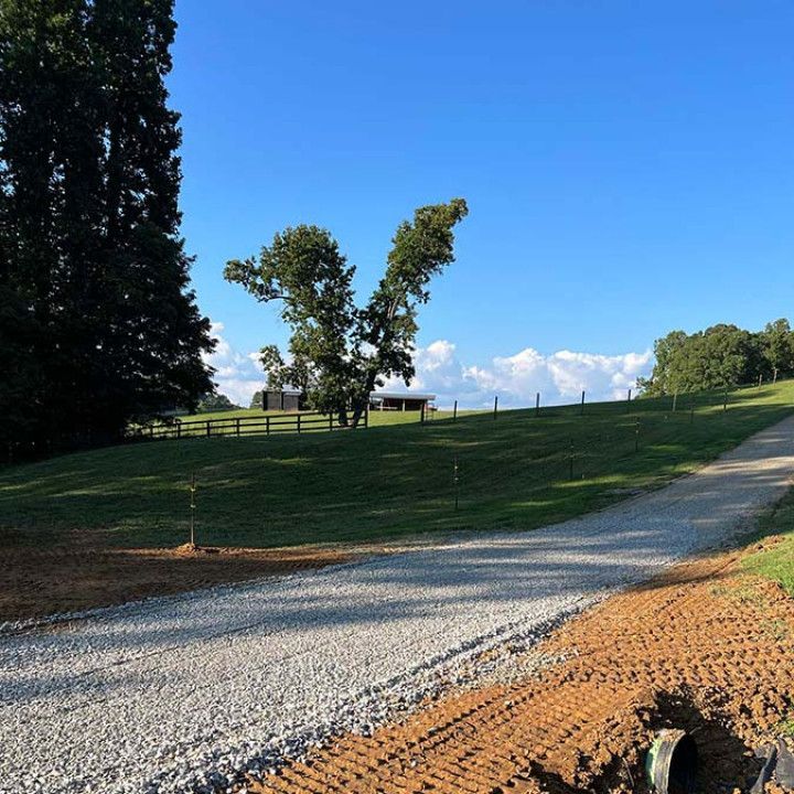 Gravel driveway leads up a grassy hill towards a fence and trees under a blue sky.