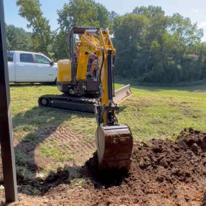 Yellow excavator digging a trench in a grassy field on a sunny day. White truck in the background.
