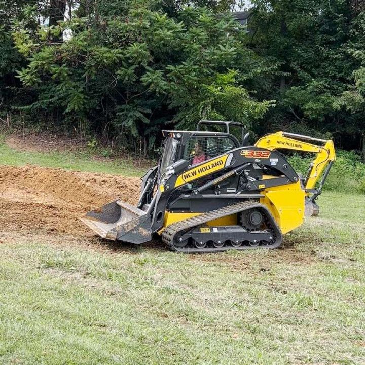 Yellow and black New Holland track skid steer operating on a grassy field, digging dirt.