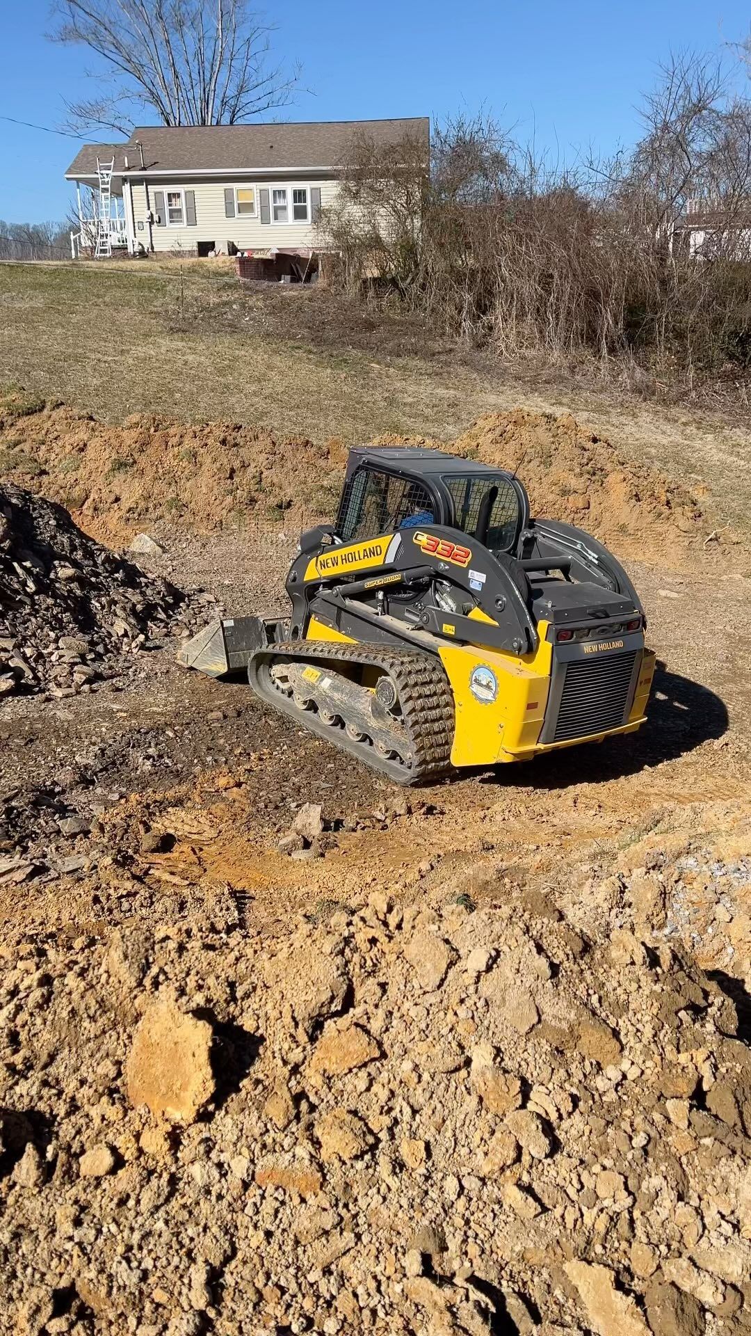 Yellow skid steer loader on a construction site, digging into the dirt. A house sits in the background.