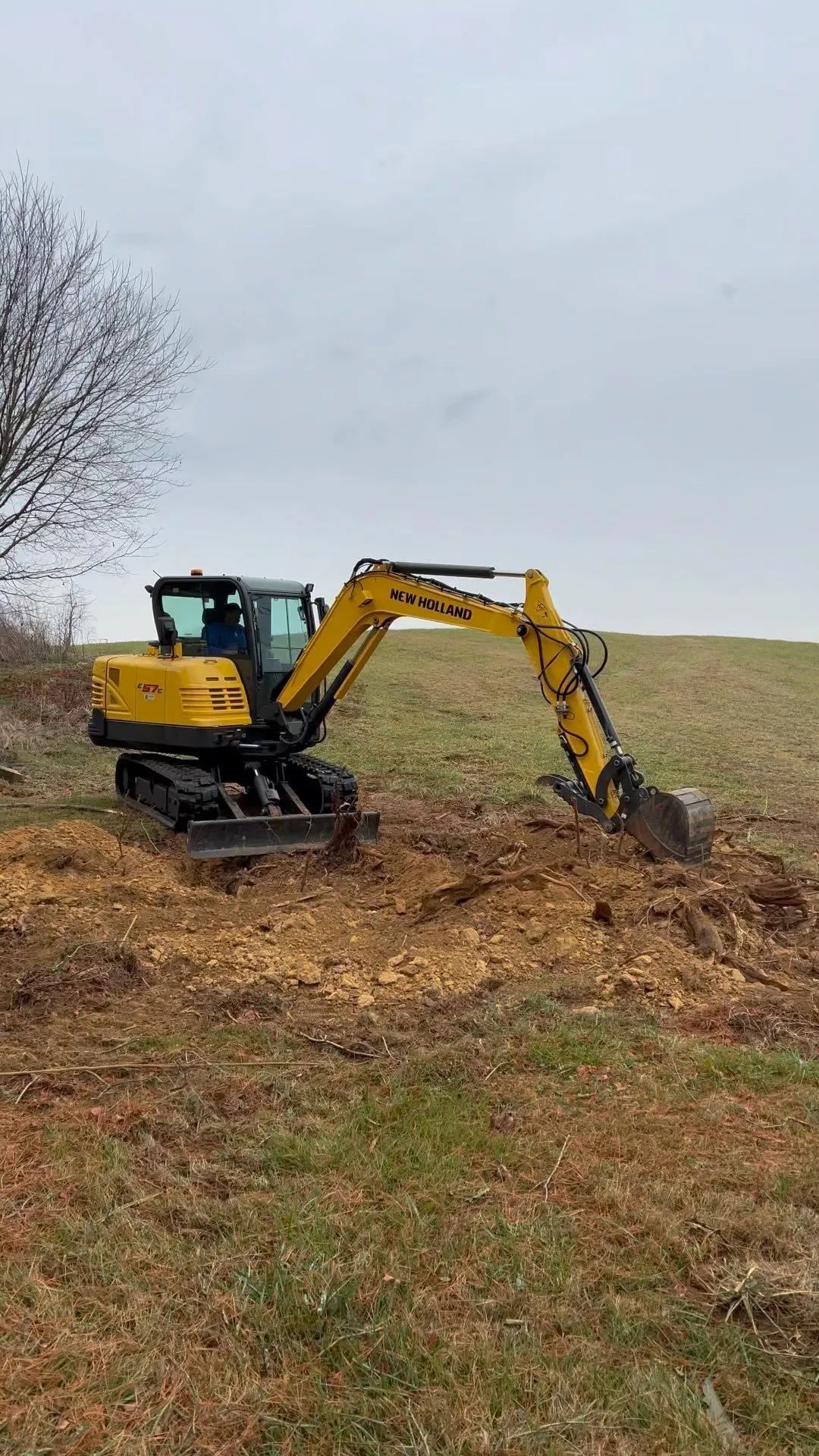 Yellow excavator digging into a dirt mound in a grassy field under an overcast sky