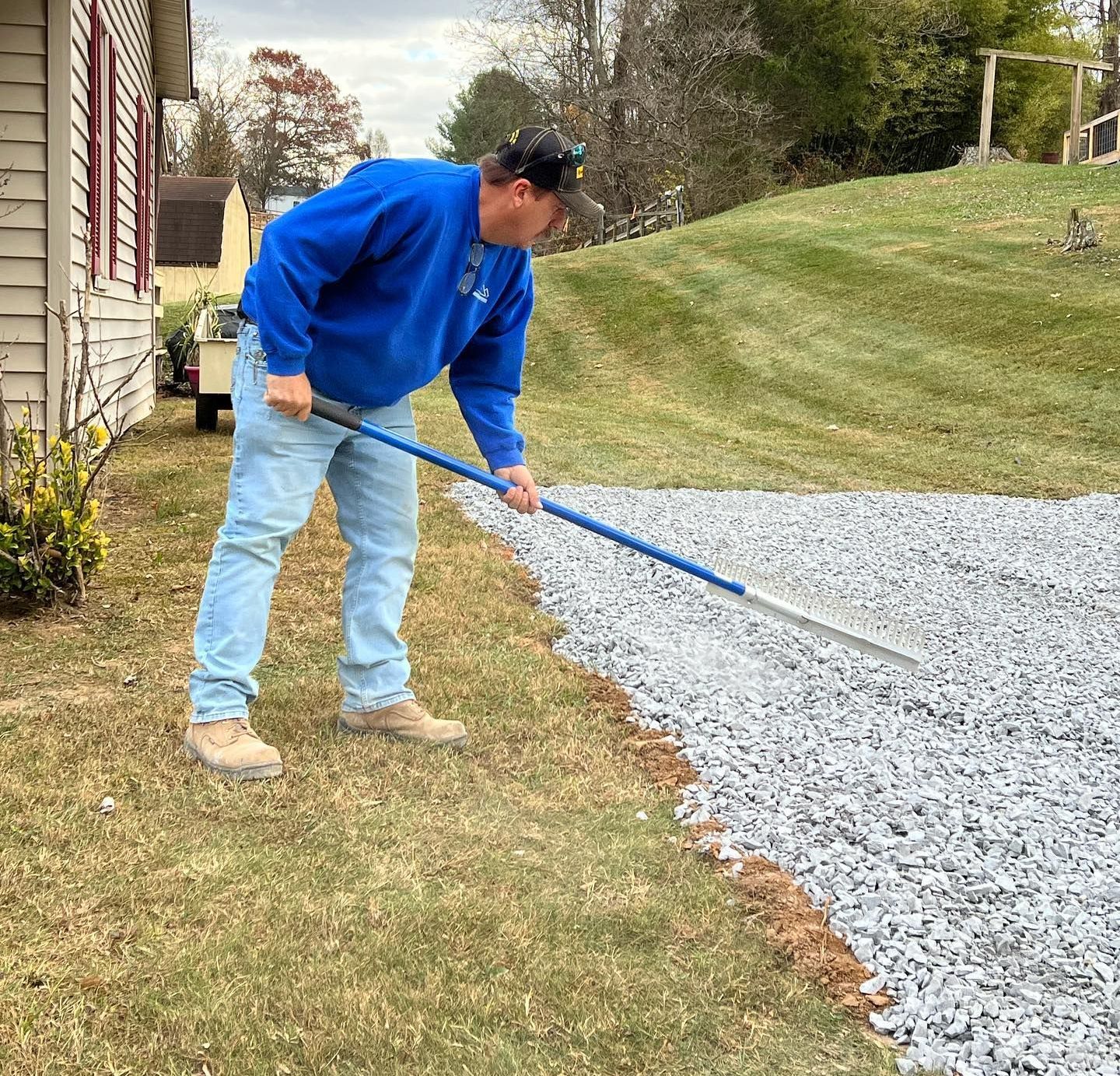 Man in blue sweatshirt and jeans raking gravel driveway beside a house and grassy hill.