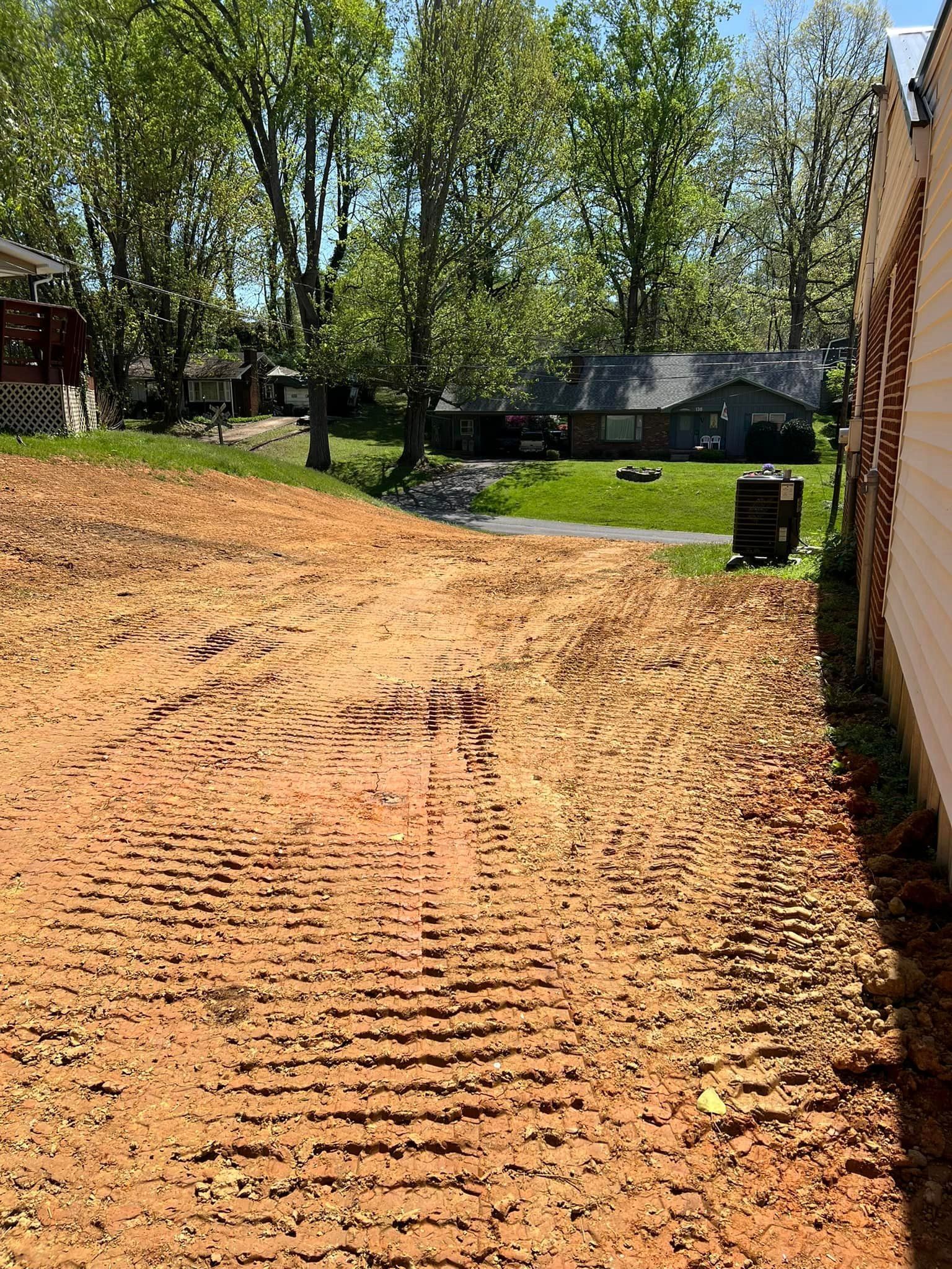 An empty, cleared dirt lot with tire tracks, next to a brick building and across from trees and houses.