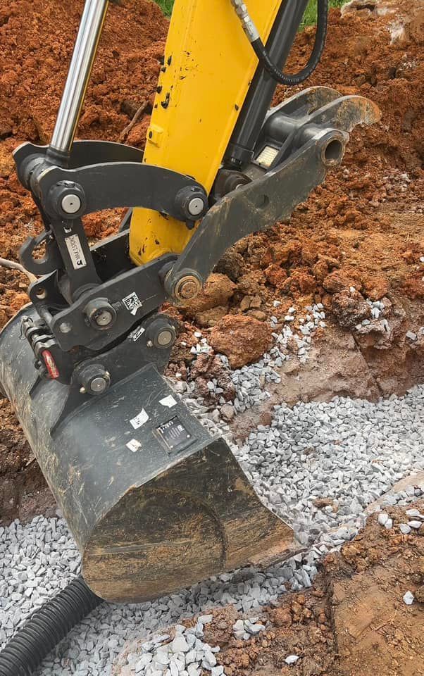 Yellow excavator bucket excavating gravel next to a buried drainage pipe.