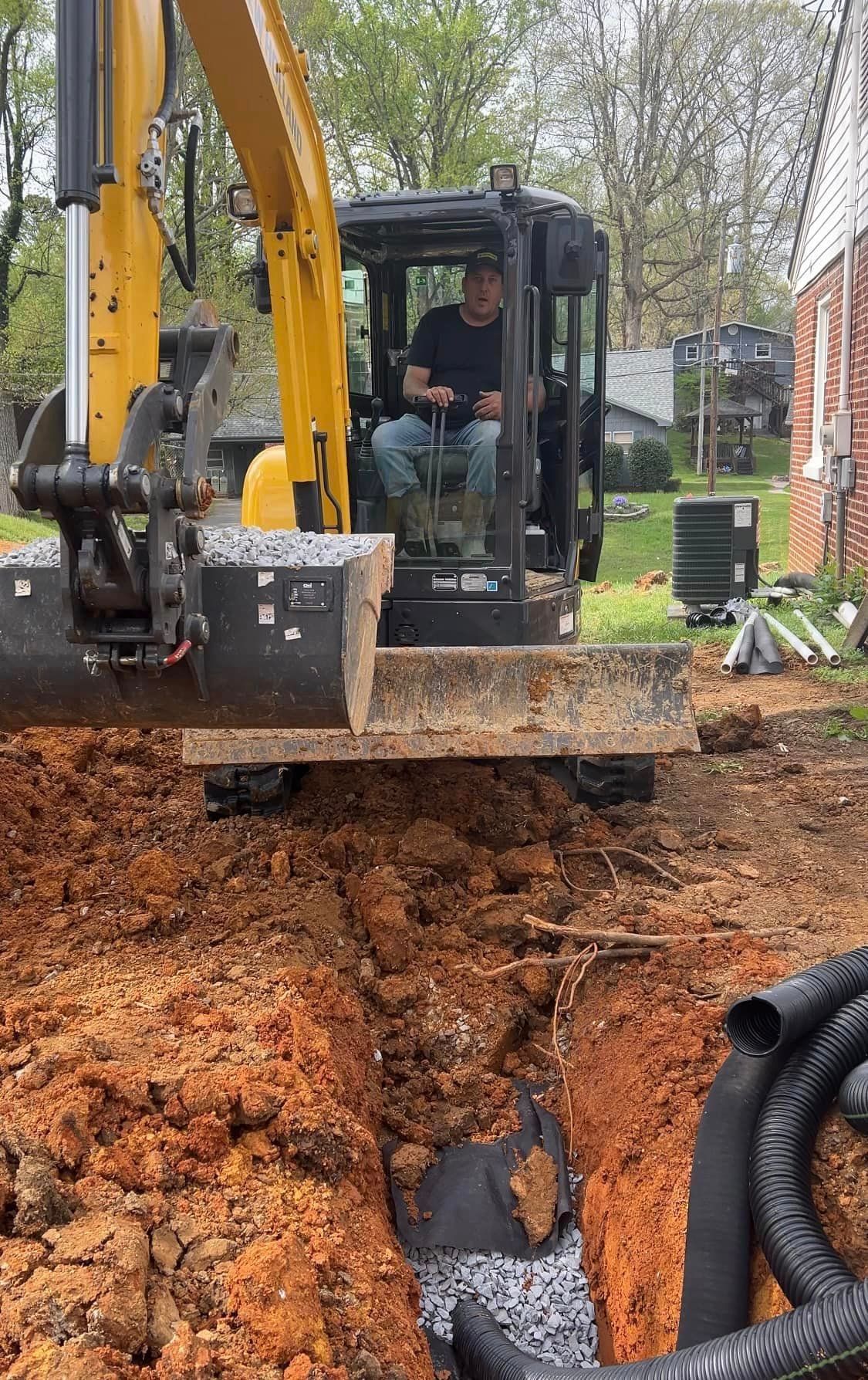 Man operating a yellow excavator digging a trench, with black drain pipe in the ground.