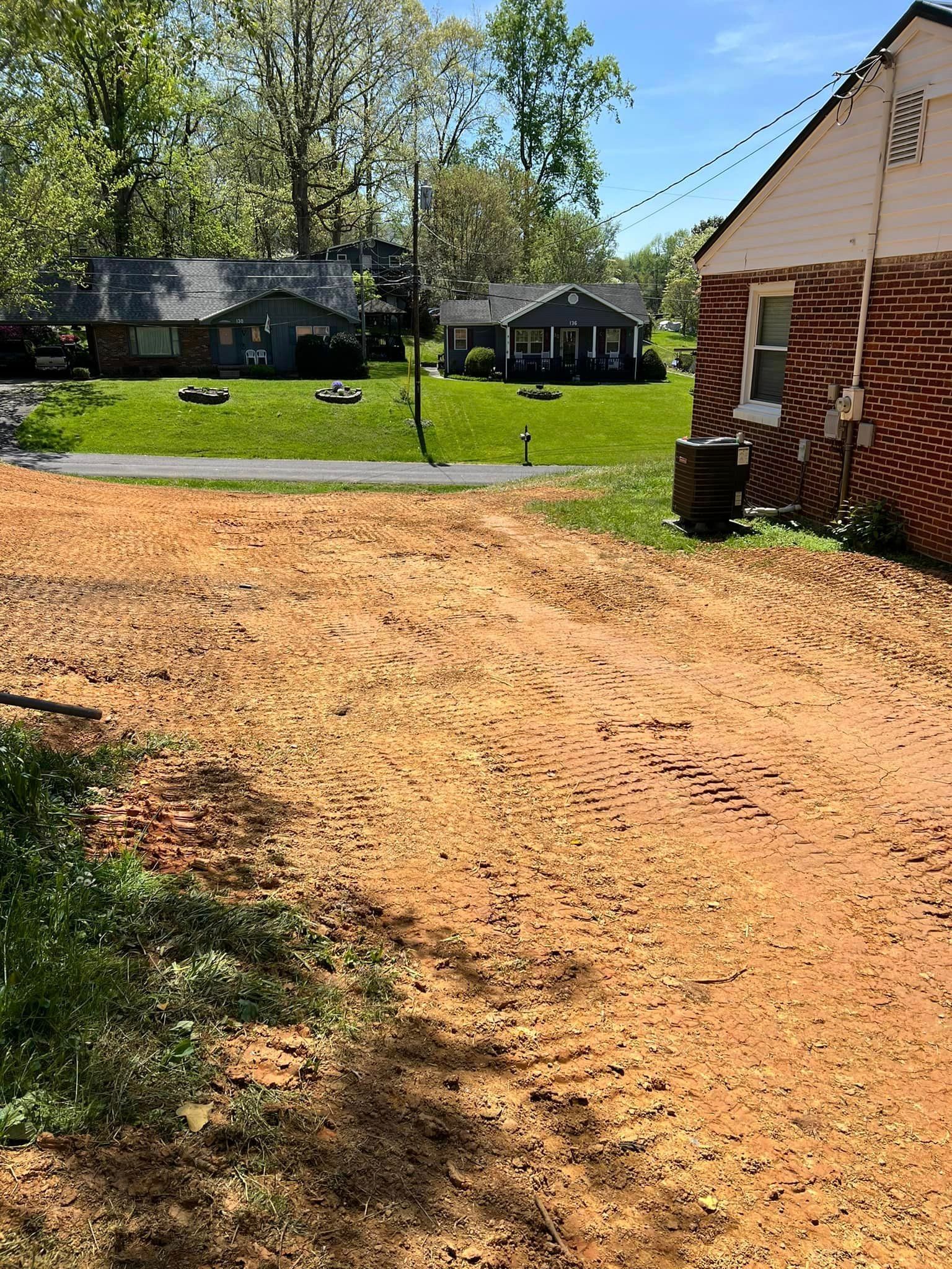 Red dirt driveway leads to grassy area with houses in the background; brick building on the right.