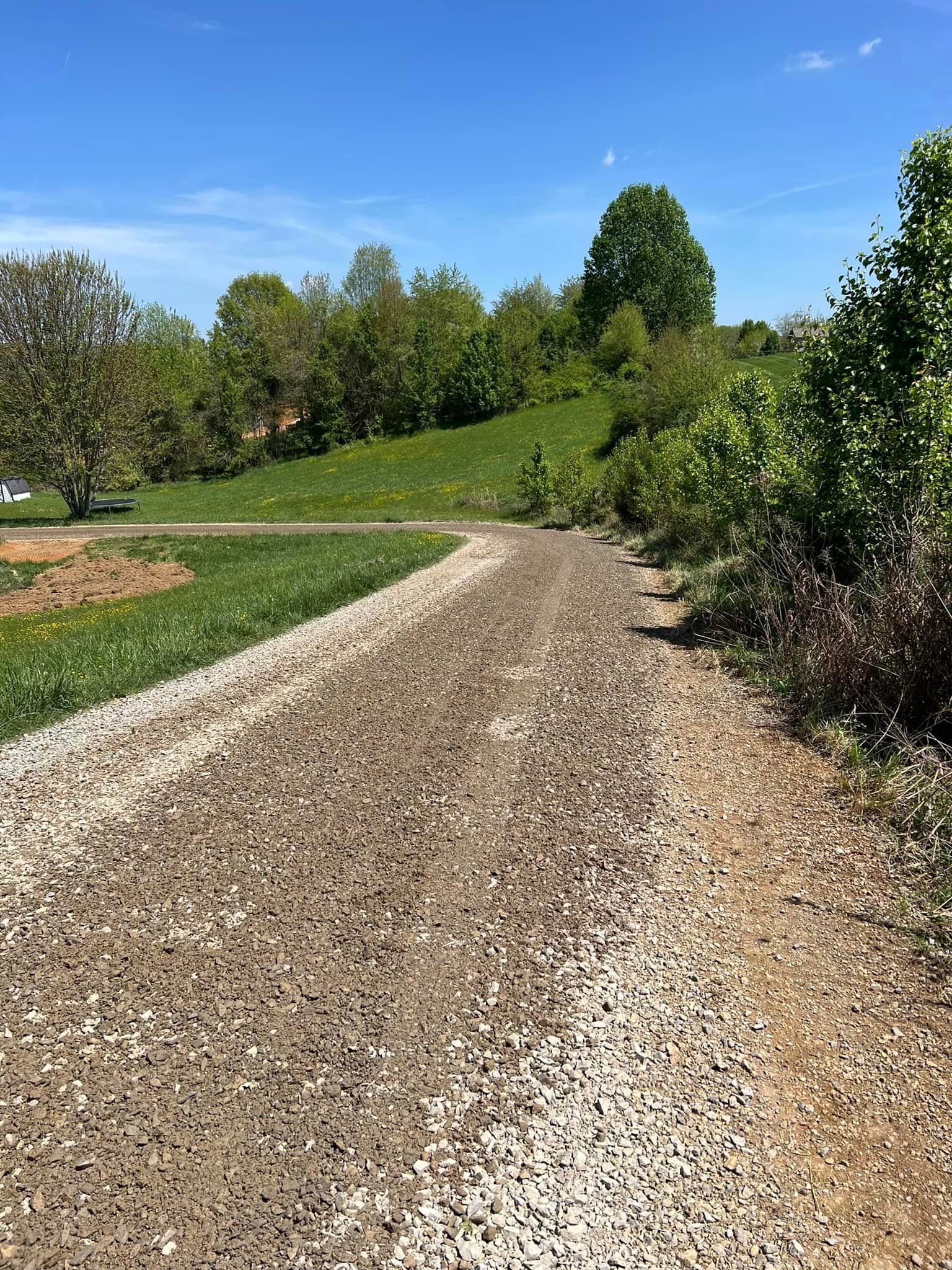 Gravel path curves uphill, flanked by green grass and trees, under a blue sky.