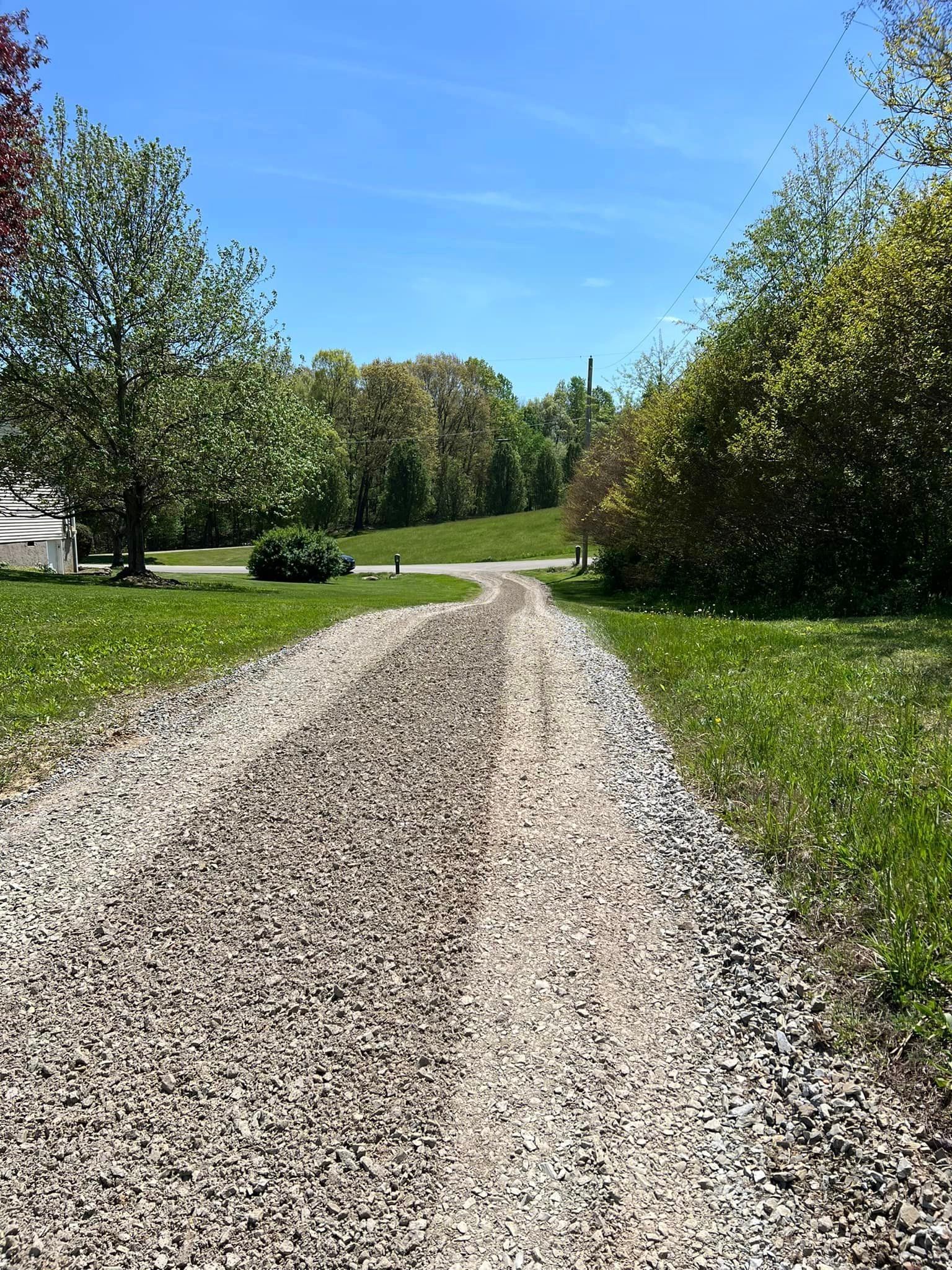 Gravel driveway curves uphill, flanked by green grass and trees, under a bright blue sky.