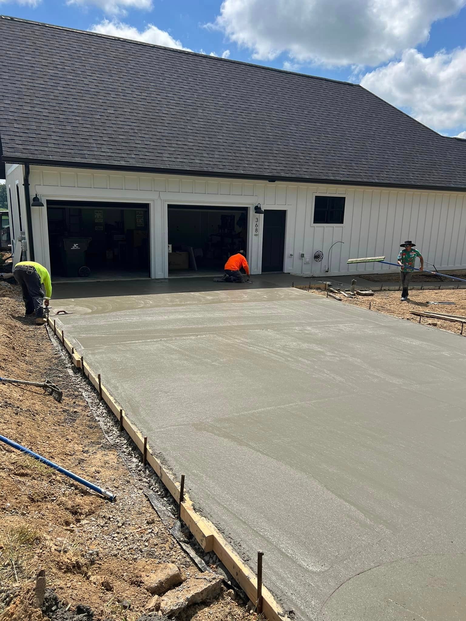Construction workers pouring concrete driveway in front of a house with black roof, on a sunny day.