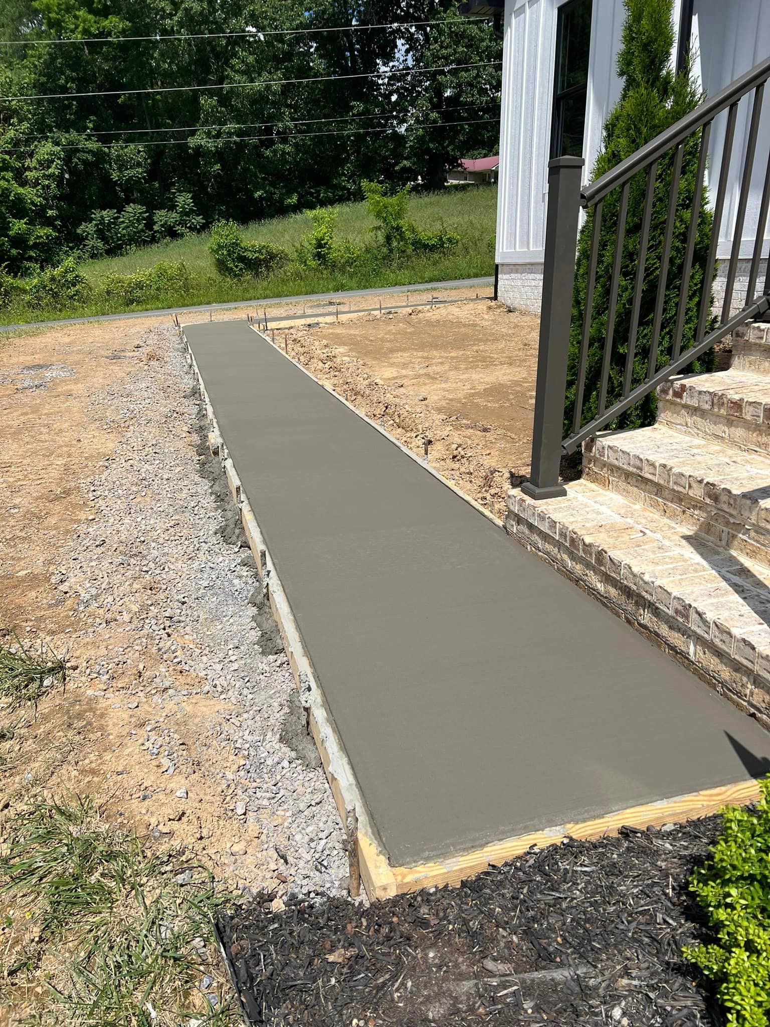 Freshly poured concrete walkway leading to house steps. Edged with gravel and soil, steps have wrought iron railing.