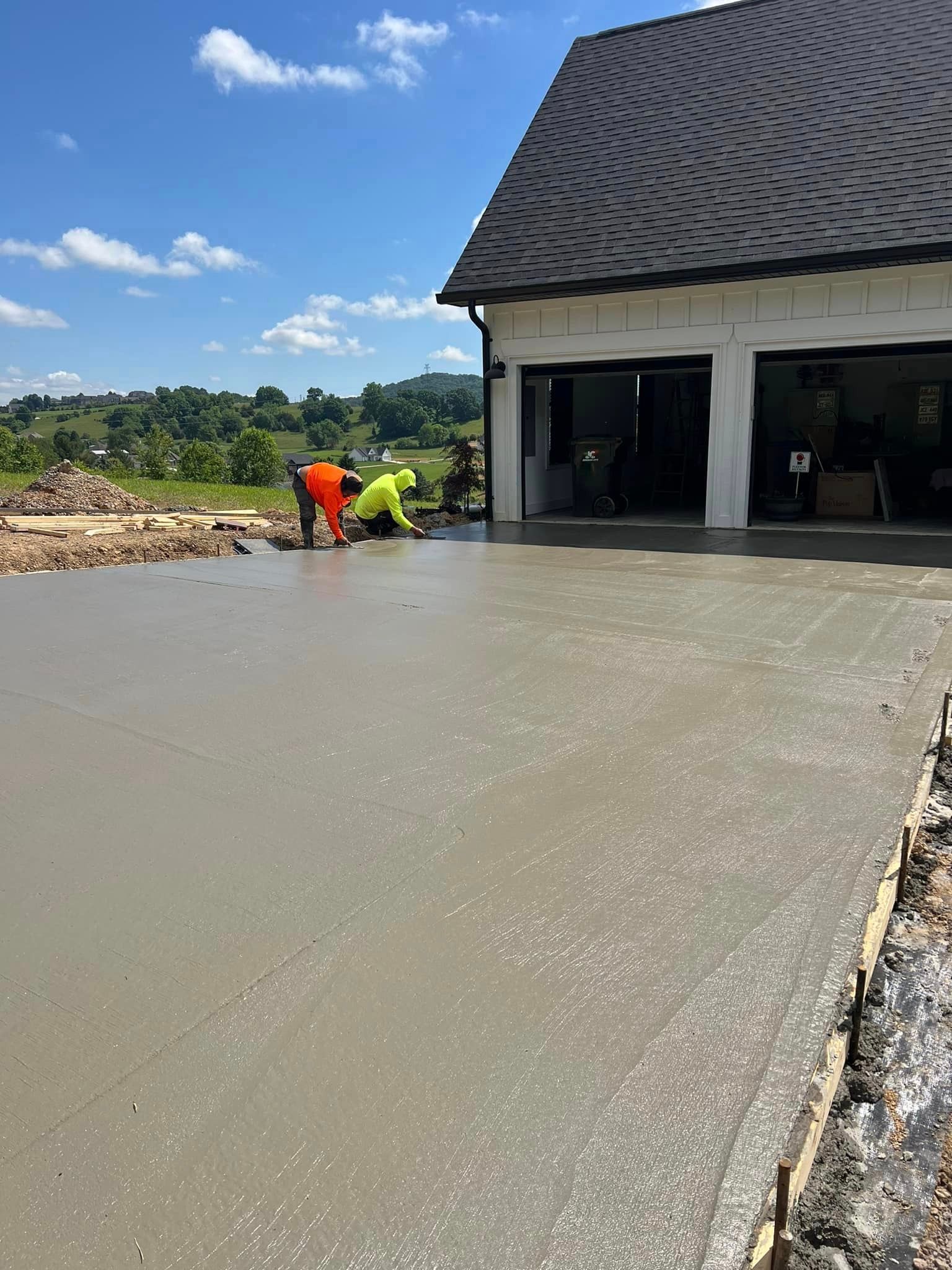 Workers in safety vests finishing concrete driveway in front of a house on a sunny day.