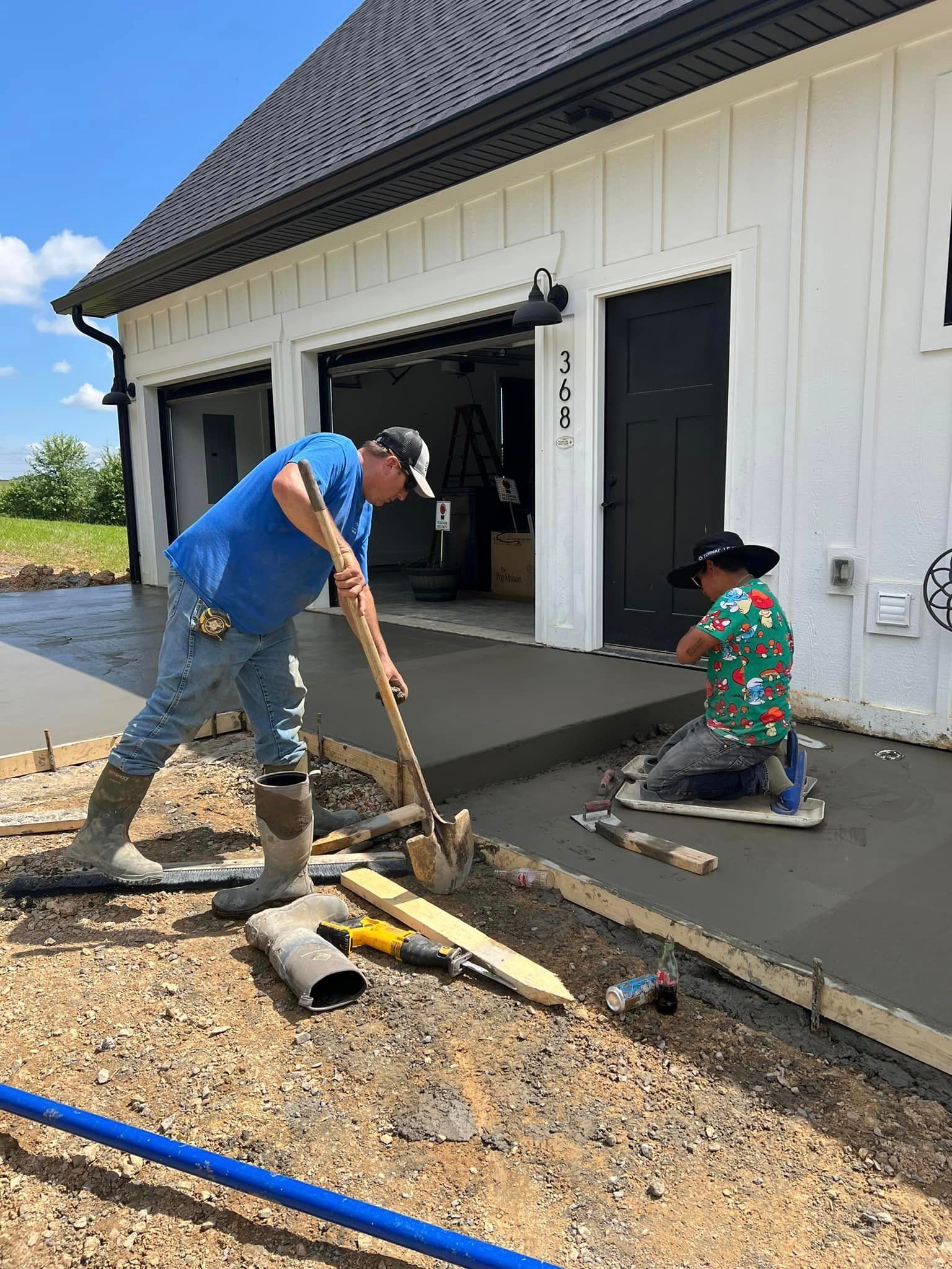 Two people laying concrete near a white garage. One shovels, the other kneels and works with tools.