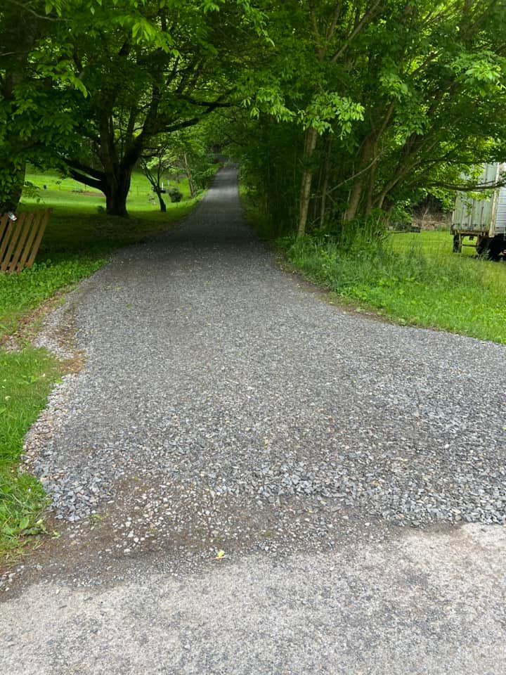 Gravel driveway leading uphill, lined with green trees and grass, with a glimpse of sky at the top.