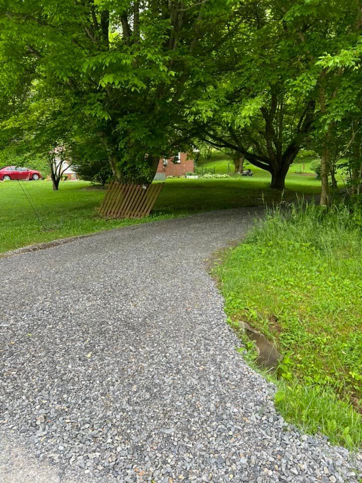 Gravel driveway leading to a house, flanked by green grass and trees. A red car is visible in the distance.
