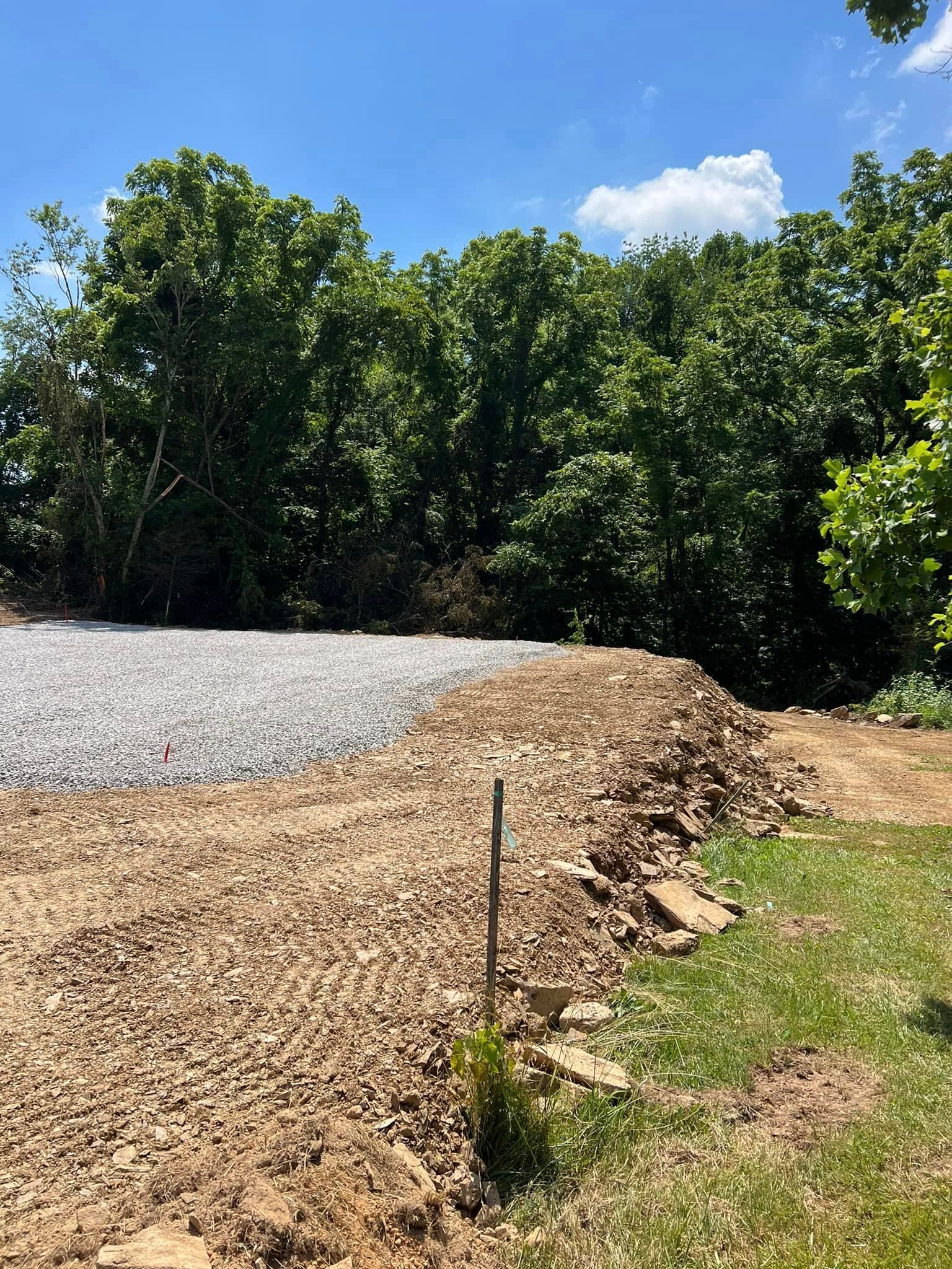 A constructed dirt and gravel area, transitioning to a bed of gravel with trees in the background under a blue sky.