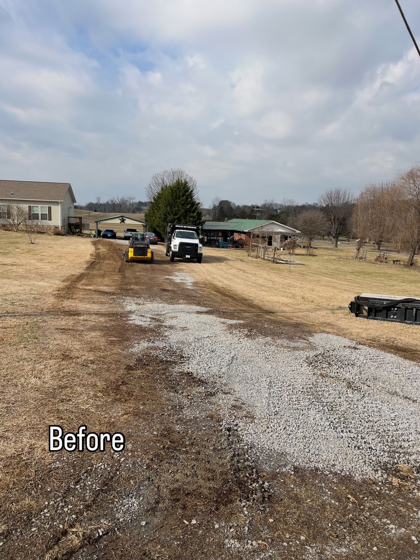 Dirt driveway with a white truck and a yellow piece of equipment. Buildings and brown grass in the background.