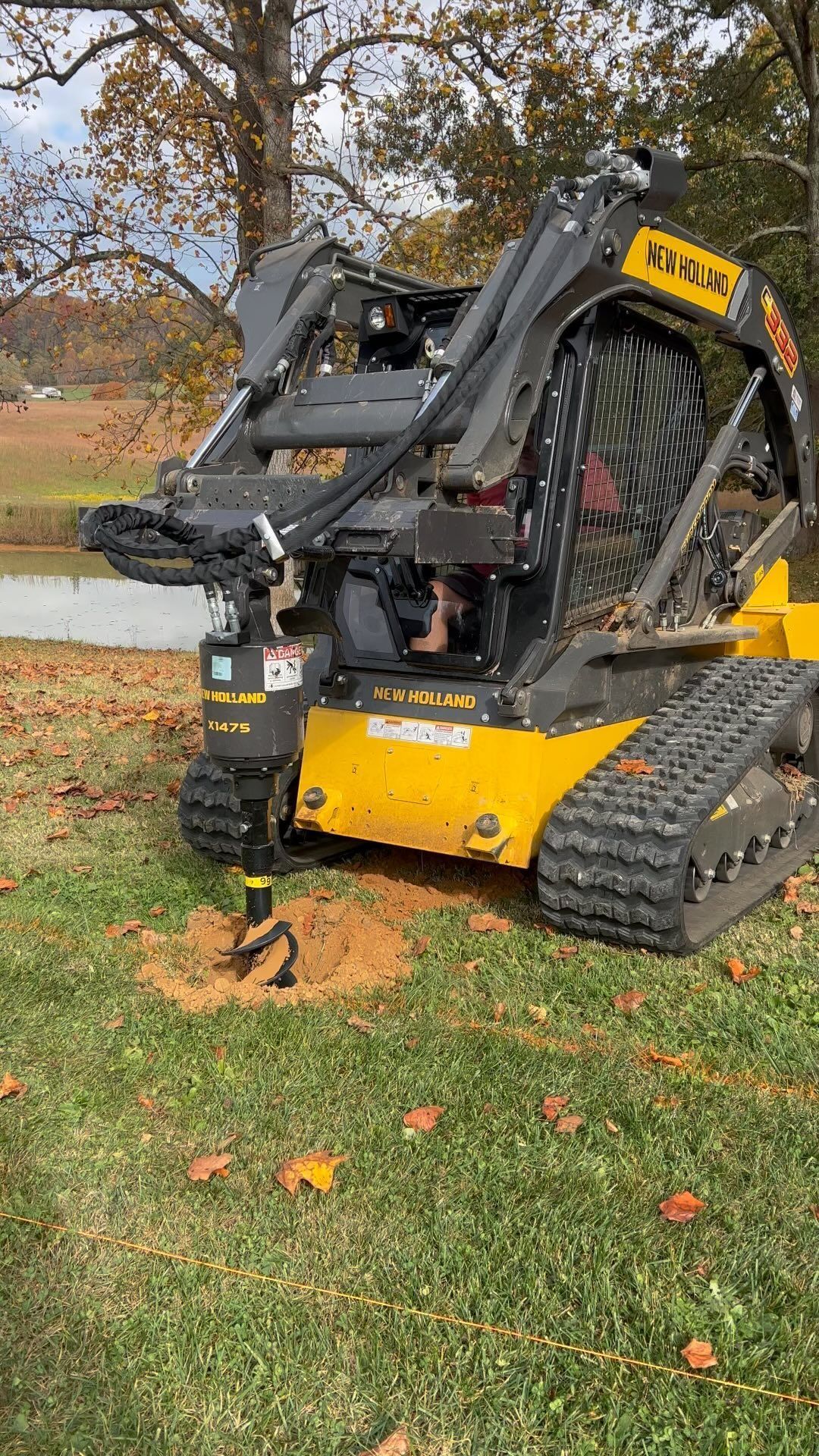 Yellow and black tracked skid steer with auger drilling into the ground, outdoors.