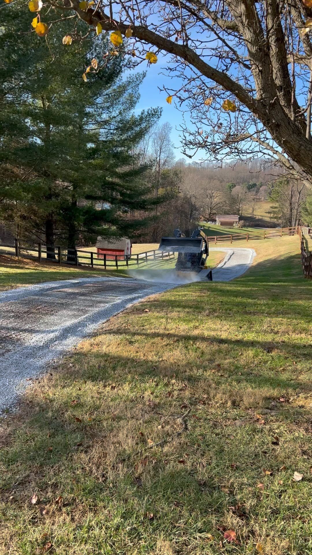 Gravel driveway with a dark truck driving away. Trees and a red and white building in the background on a sunny day.