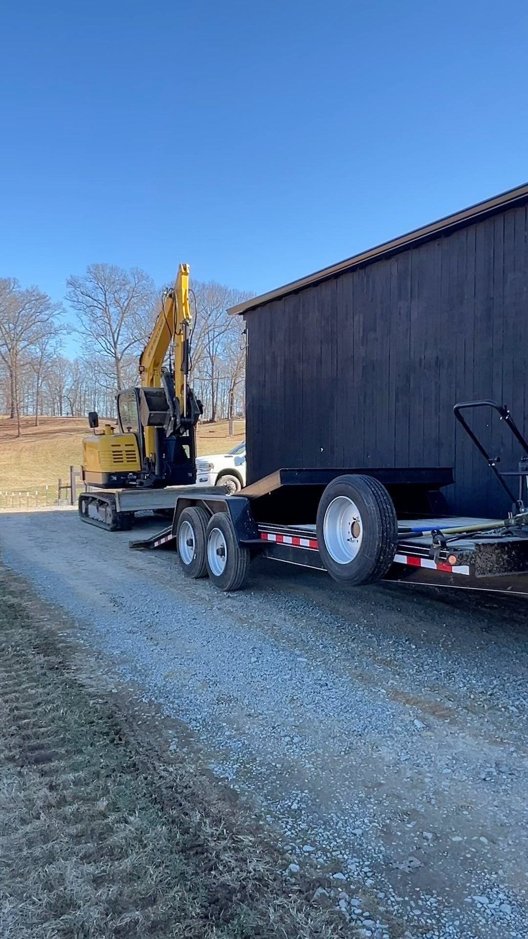 Excavator being loaded onto a trailer beside a dark building on a gravel road, sunny day.