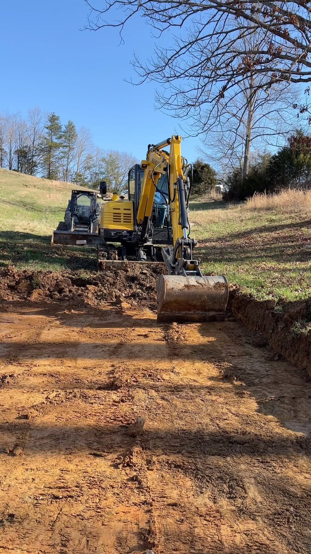 Yellow excavator digging in a dirt area, with a grassy hill in the background under a blue sky.