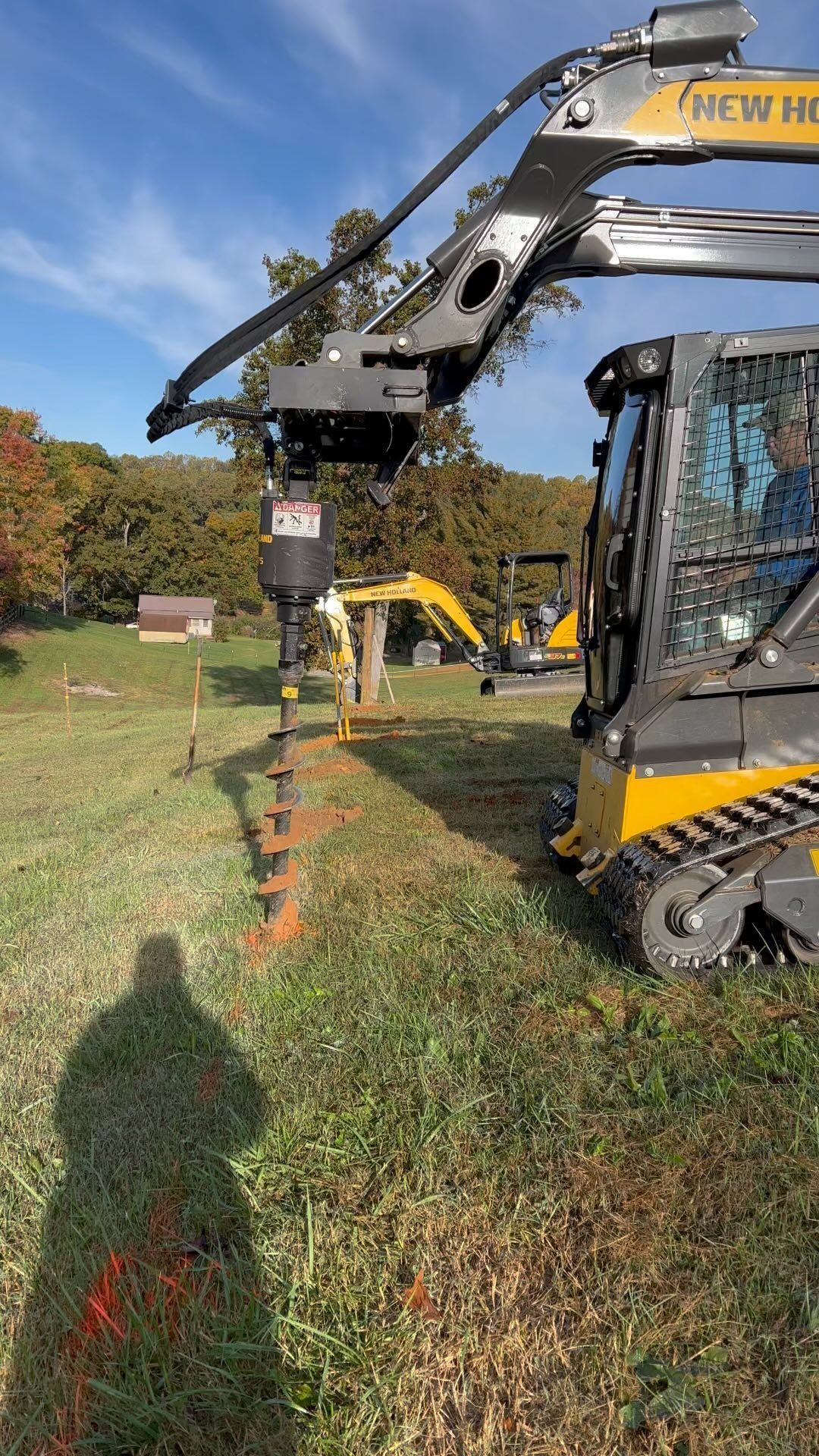 Skid steer with auger drilling holes in a line marked with orange spray paint, in grassy field.