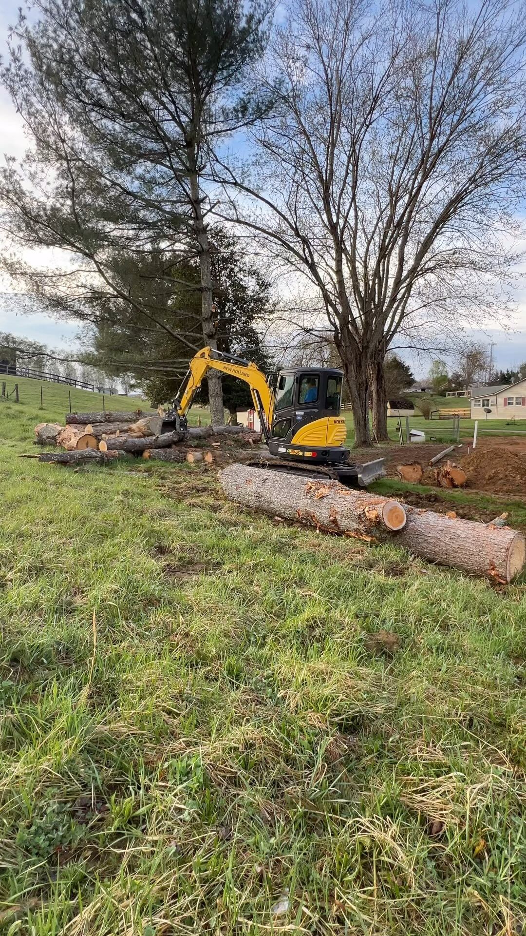 A yellow excavator moving logs in a grassy field, near trees and a fence.