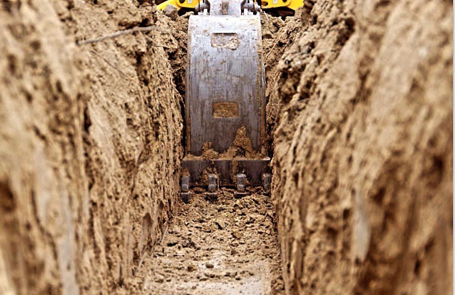 Excavator bucket in a narrow trench, digging into brown soil.