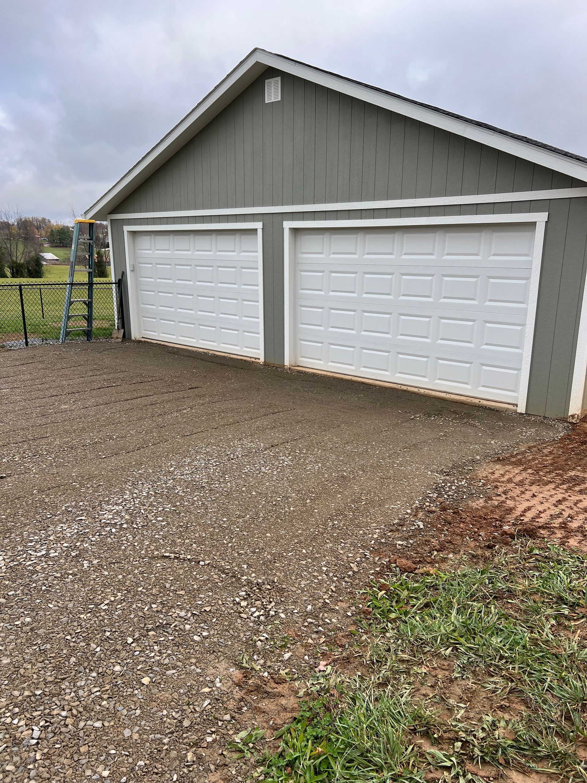 Two-car garage with white doors, green siding, gravel driveway, and overcast sky.