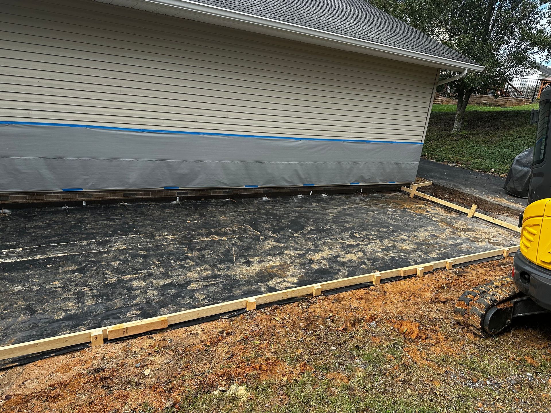 Construction site: wooden frame for a concrete patio next to a house with beige siding.