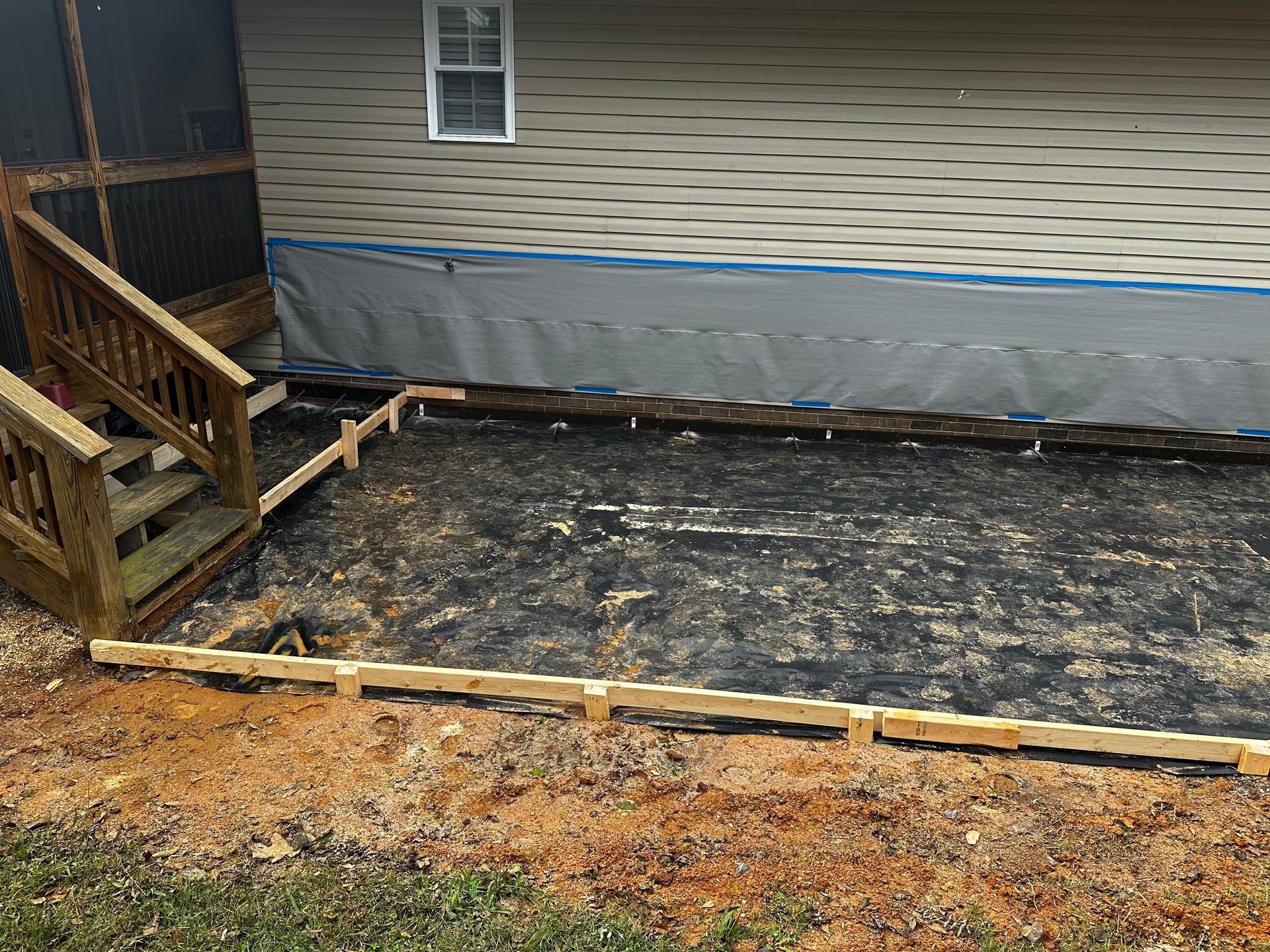 Construction site with wooden forms for a concrete patio next to a house with a deck. Black fabric covers the ground.