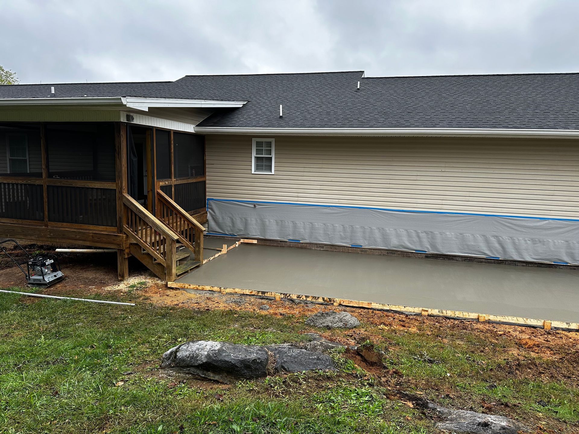 Concrete patio under house; wooden steps, screened porch to the left. Grey siding and black roof.