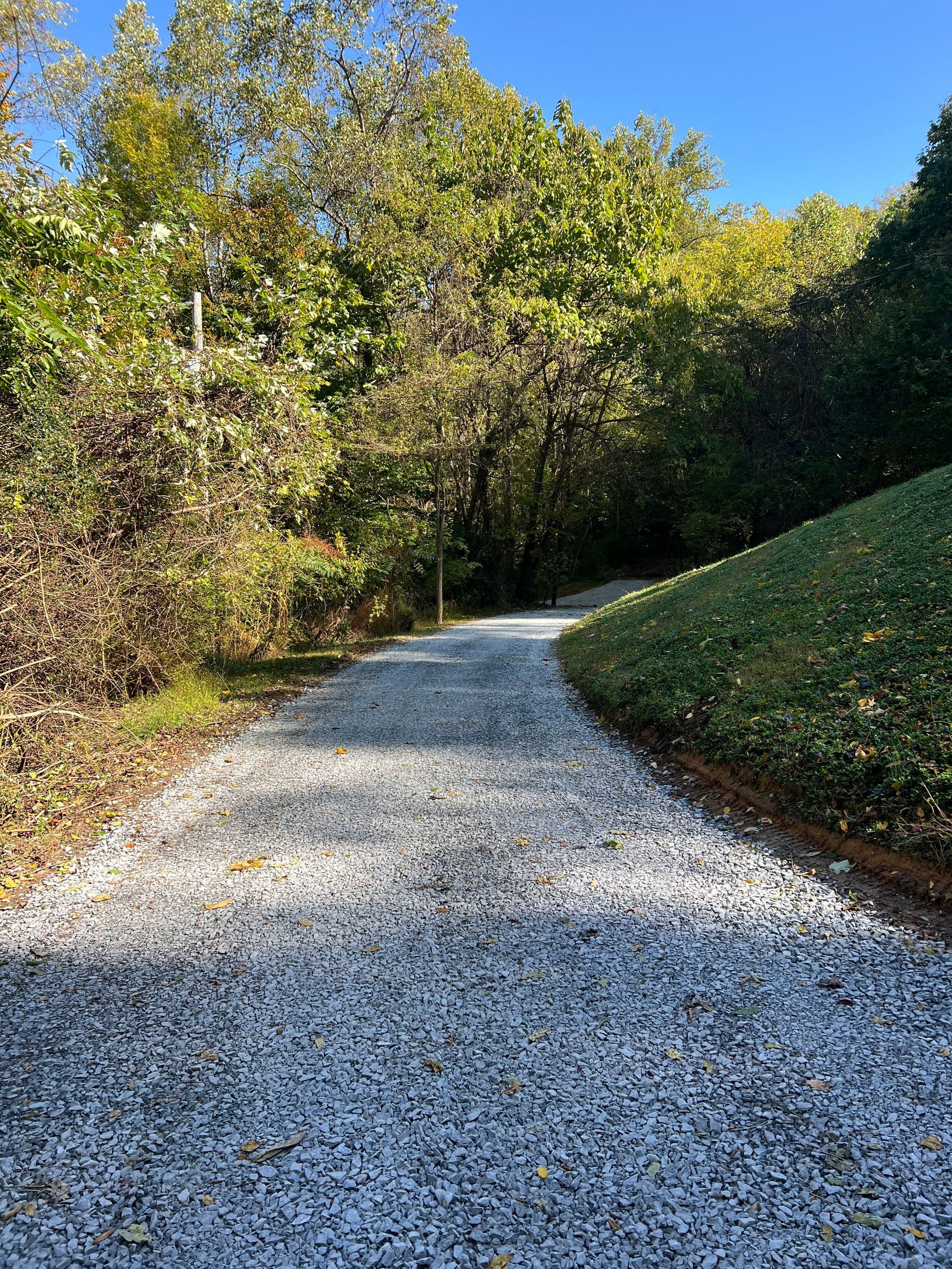 Gravel path winds uphill through a wooded area on a sunny day. Green grass and trees line the path.