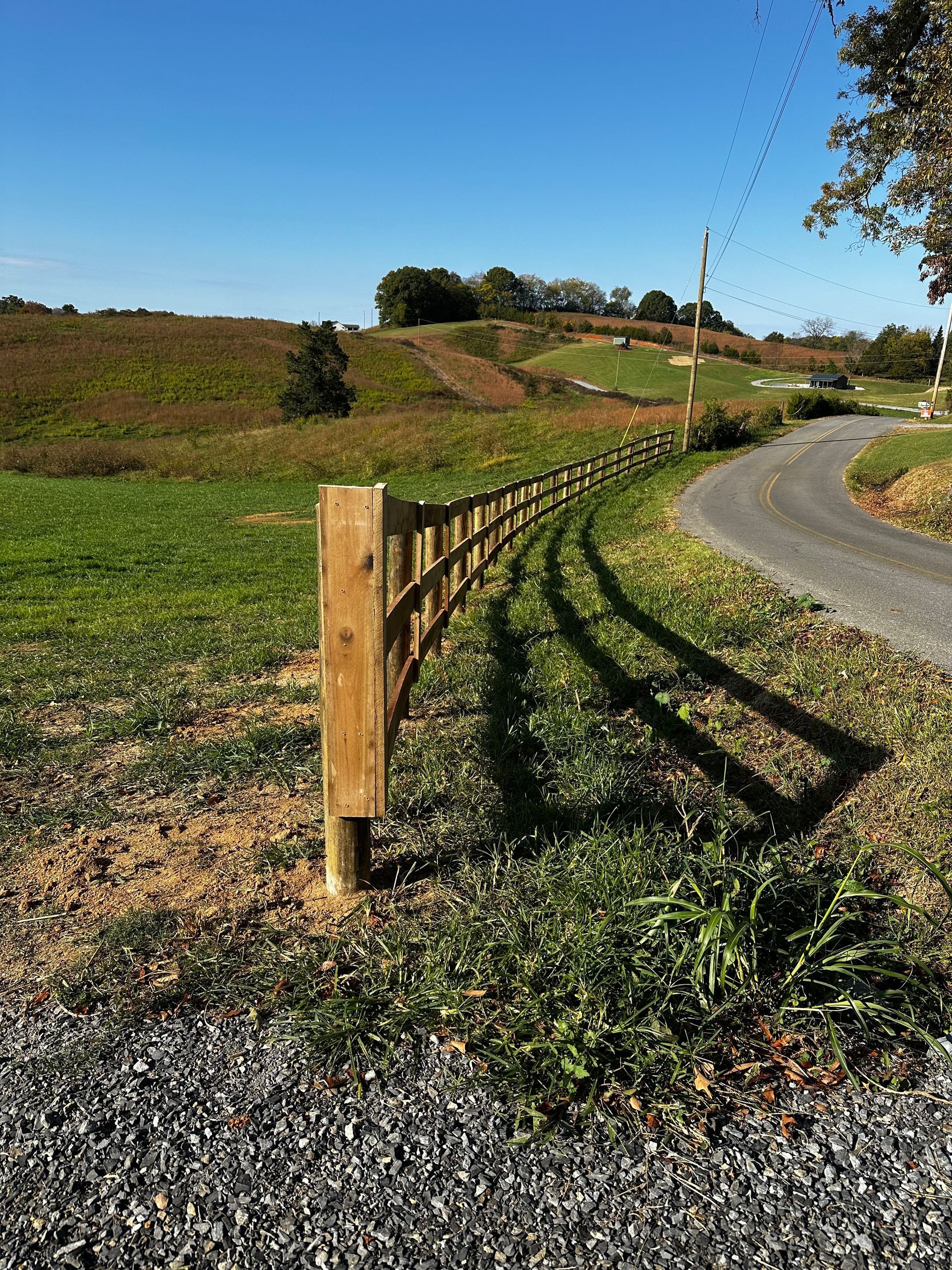 Wooden fence curving along a gravel driveway in a green field with rolling hills and a blue sky.