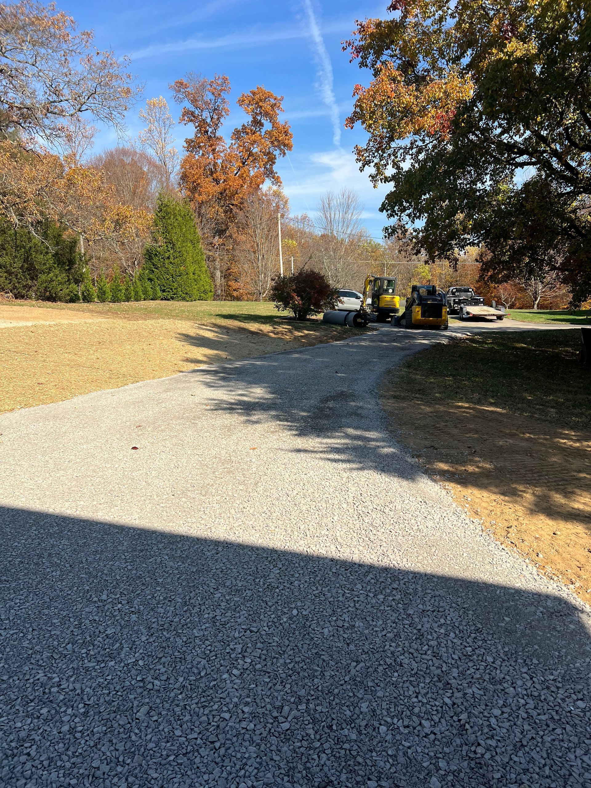 Gravel driveway leading to yard with fall foliage and construction equipment under a blue sky.