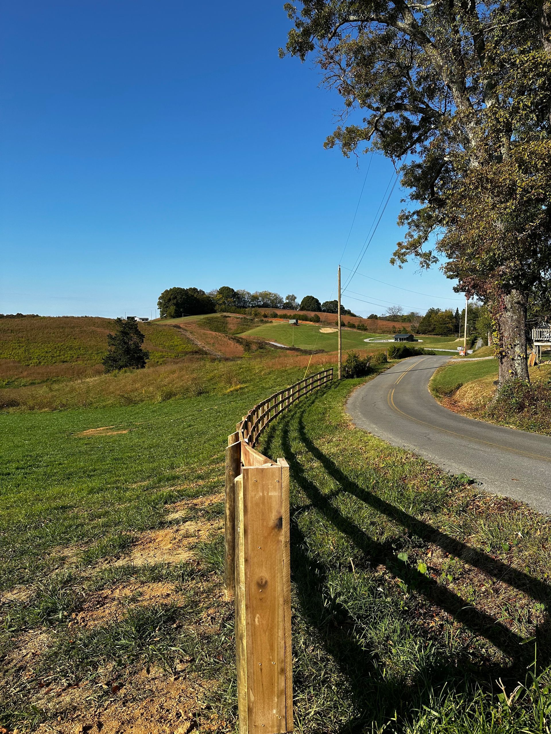 A rural scene with a curved fence, gravel road, and grassy fields under a blue sky.