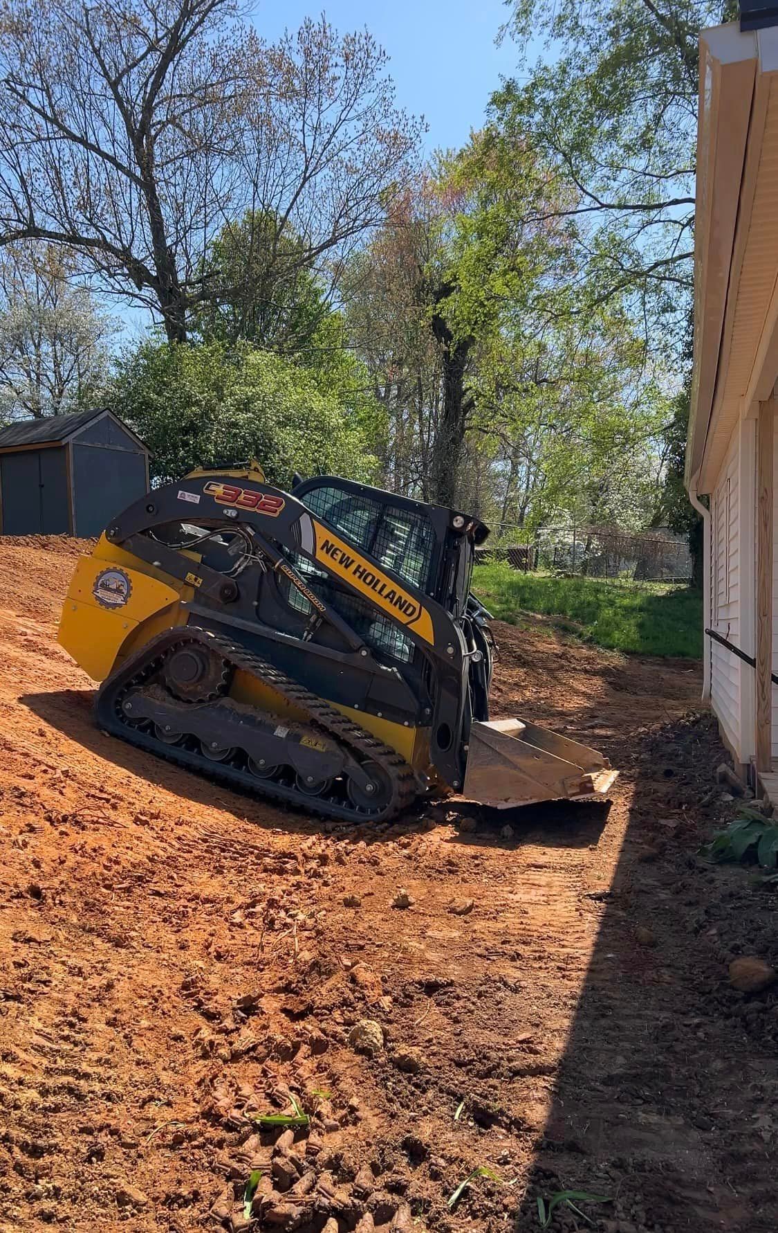 Yellow and black track loader operating near a building, in a yard with dirt and trees.