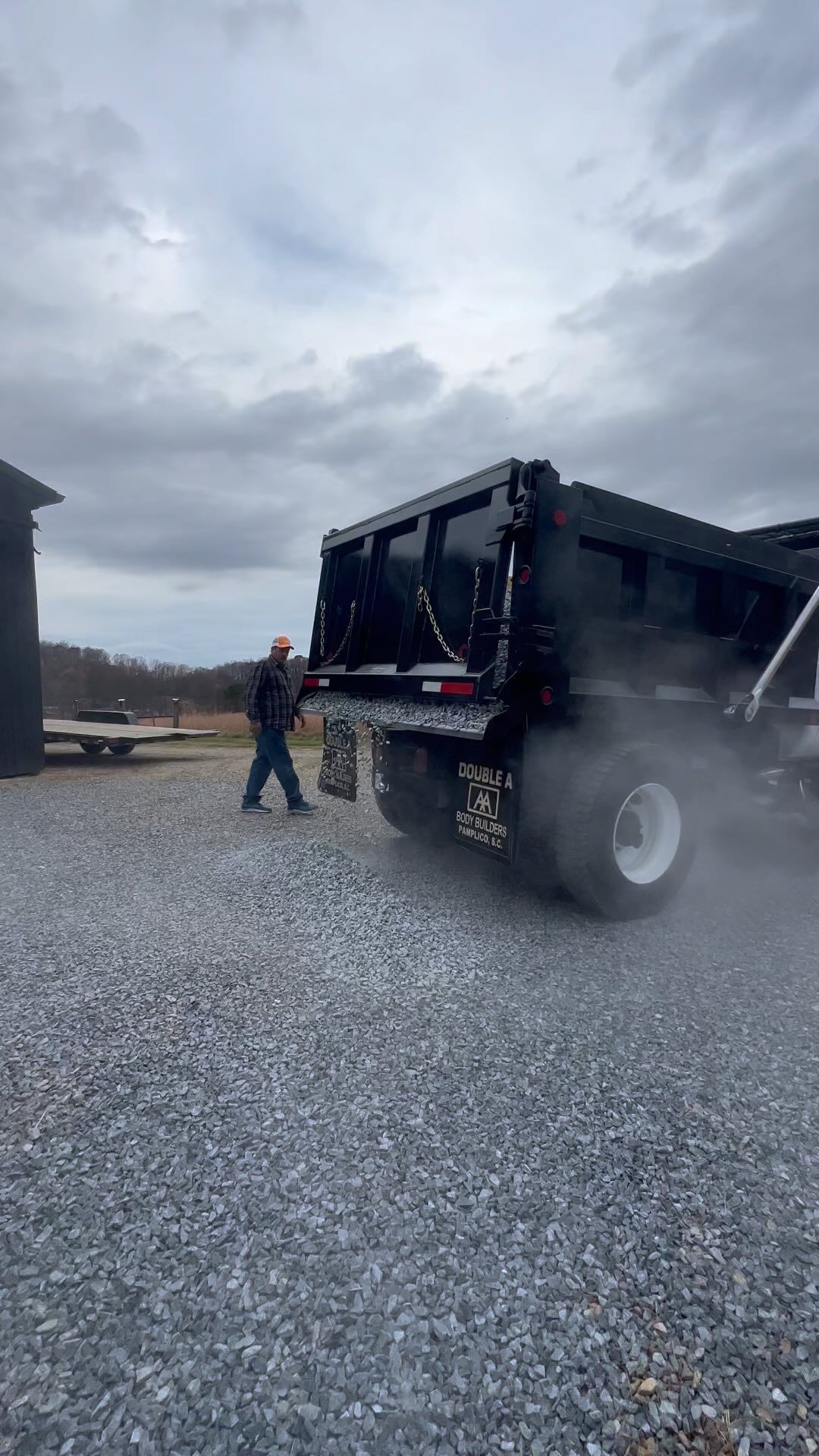 Man standing next to a black dump truck with smoke or dust rising from the tires on a gravel surface.