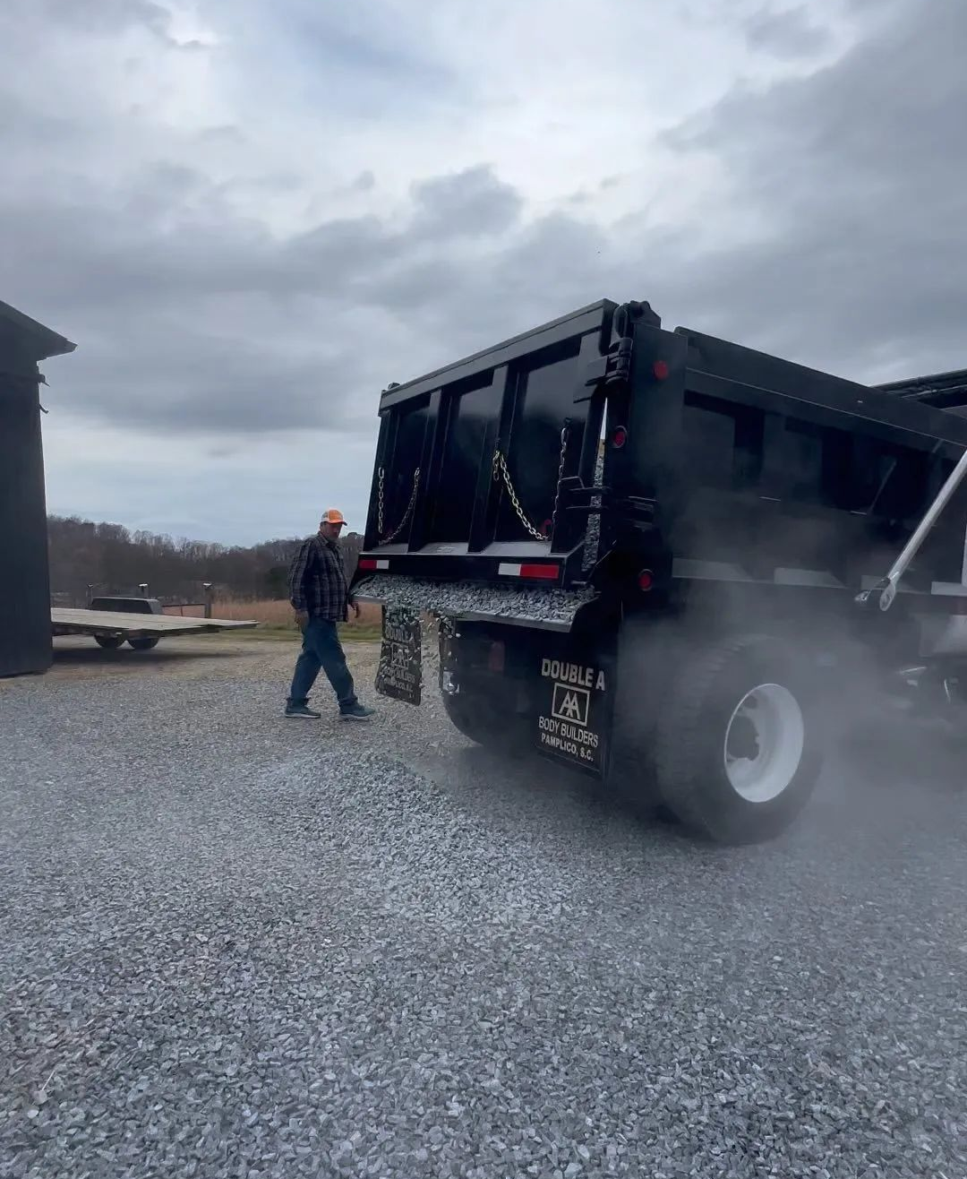 Worker standing beside a black dump truck on a gravel lot, with steam or dust rising around the rear tires.
