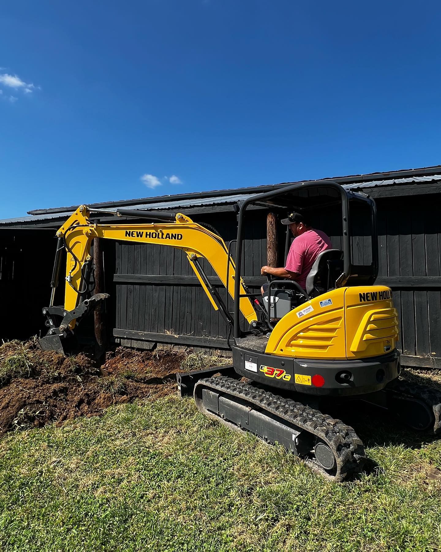 Yellow excavator digging near a black fence, operated by a person in a pink shirt on a sunny day.