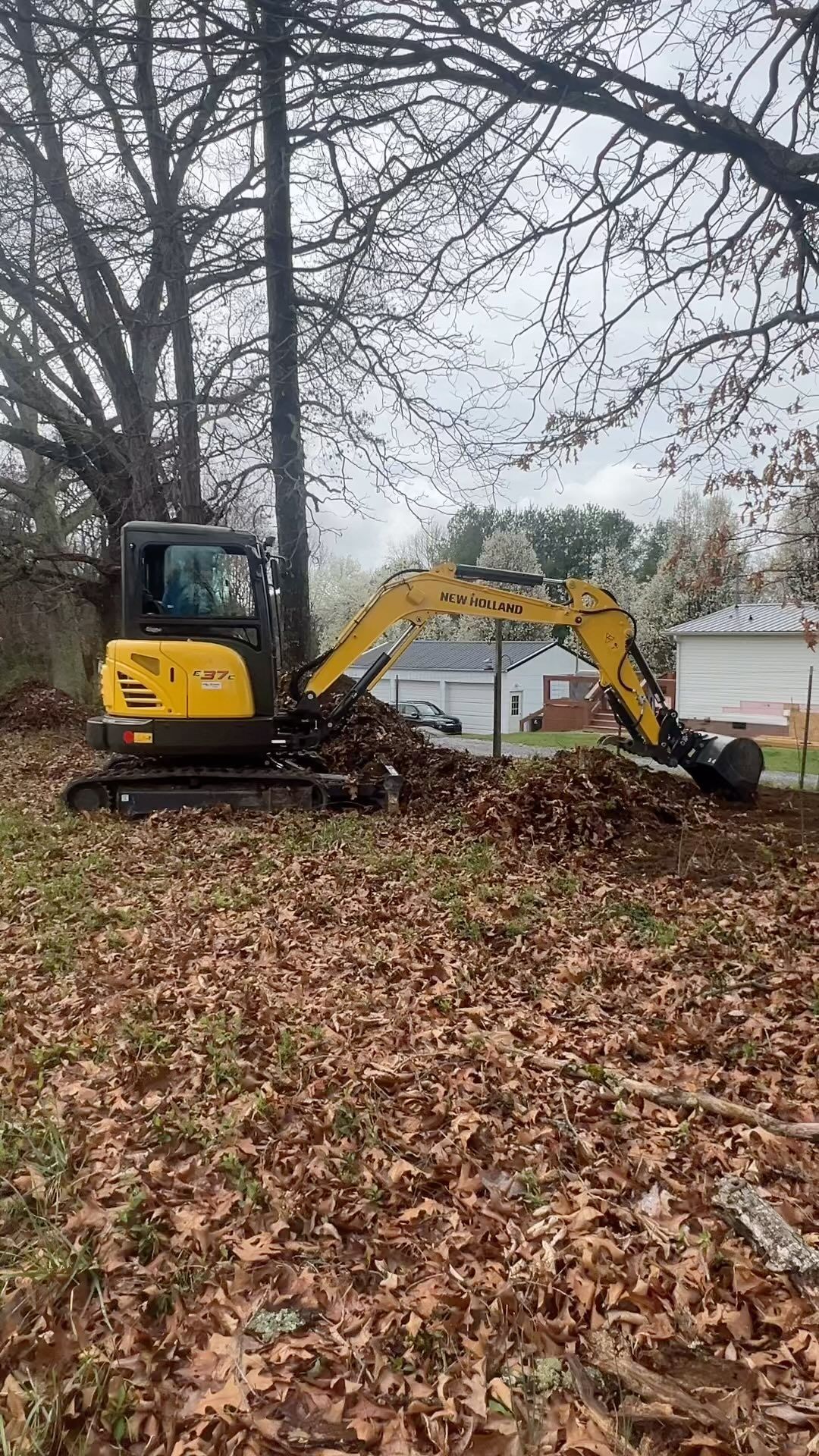 Small excavator clearing leaves near a house, trees in the background.