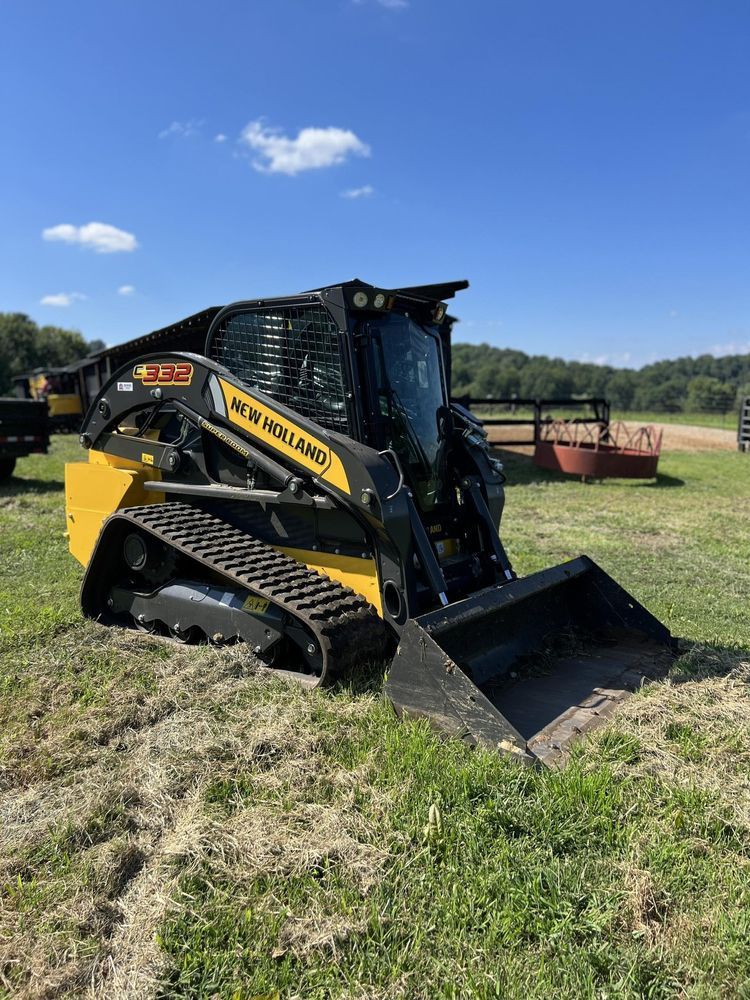 Yellow and black track loader with bucket in a grassy field on a sunny day.
