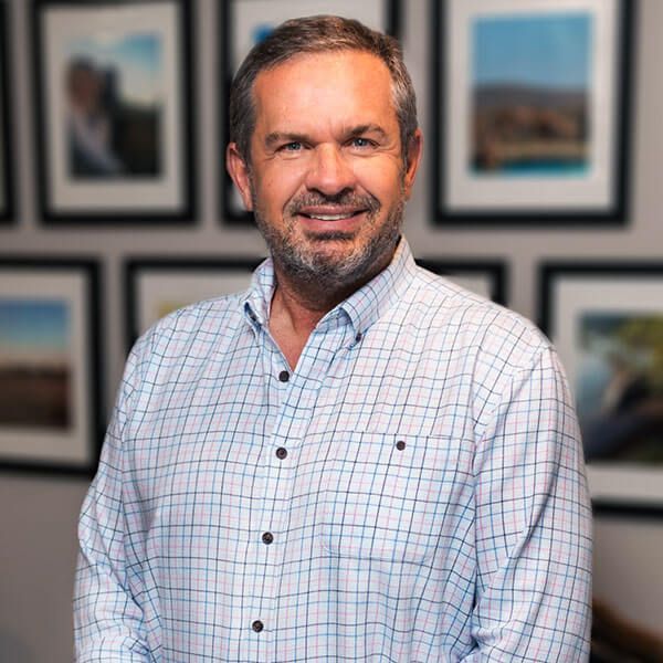 Smiling man in a plaid shirt stands in front of framed art on a wall.