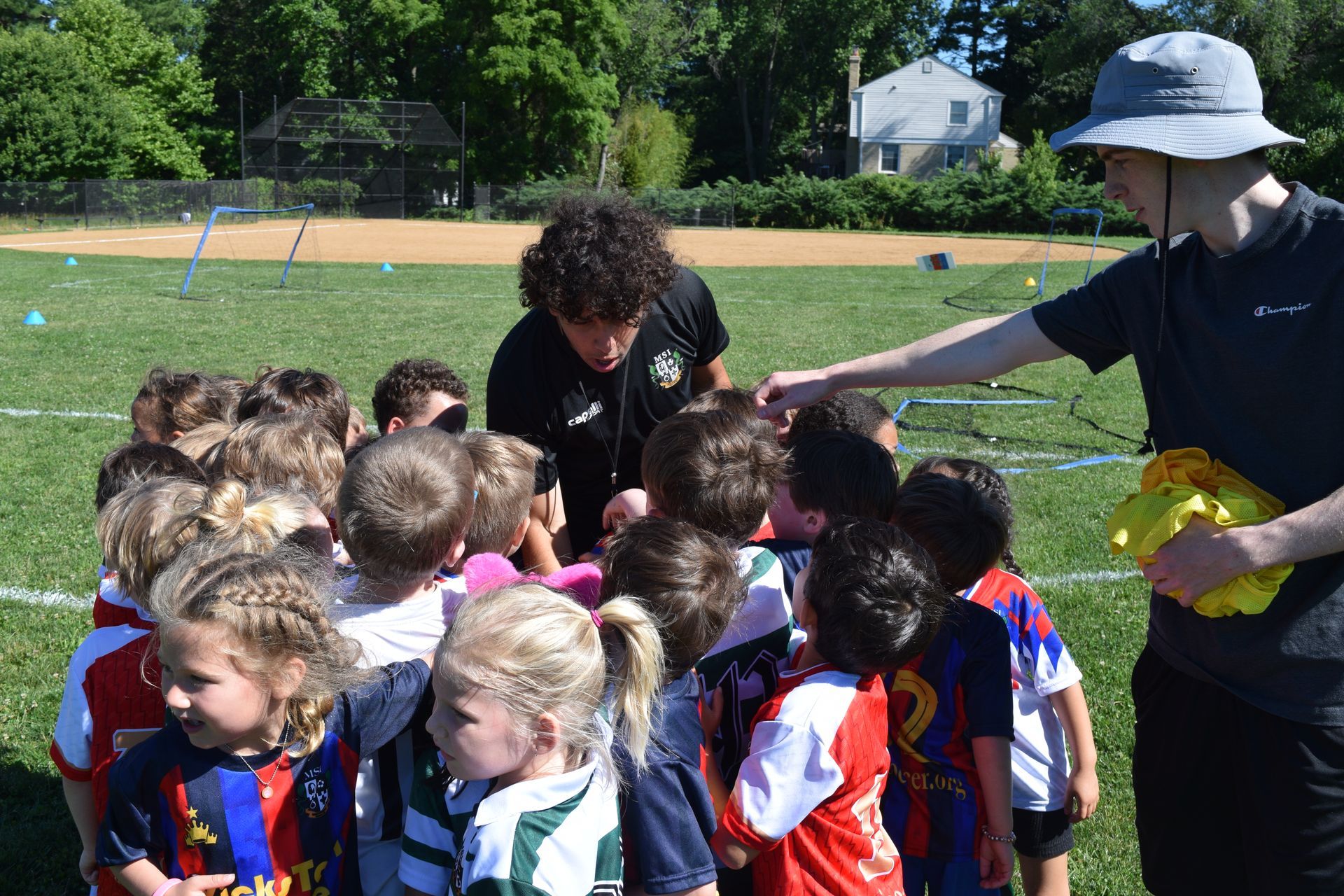 Young MSI soccer players huddle around their coach on sunny field as assistant hands out training gear during practice