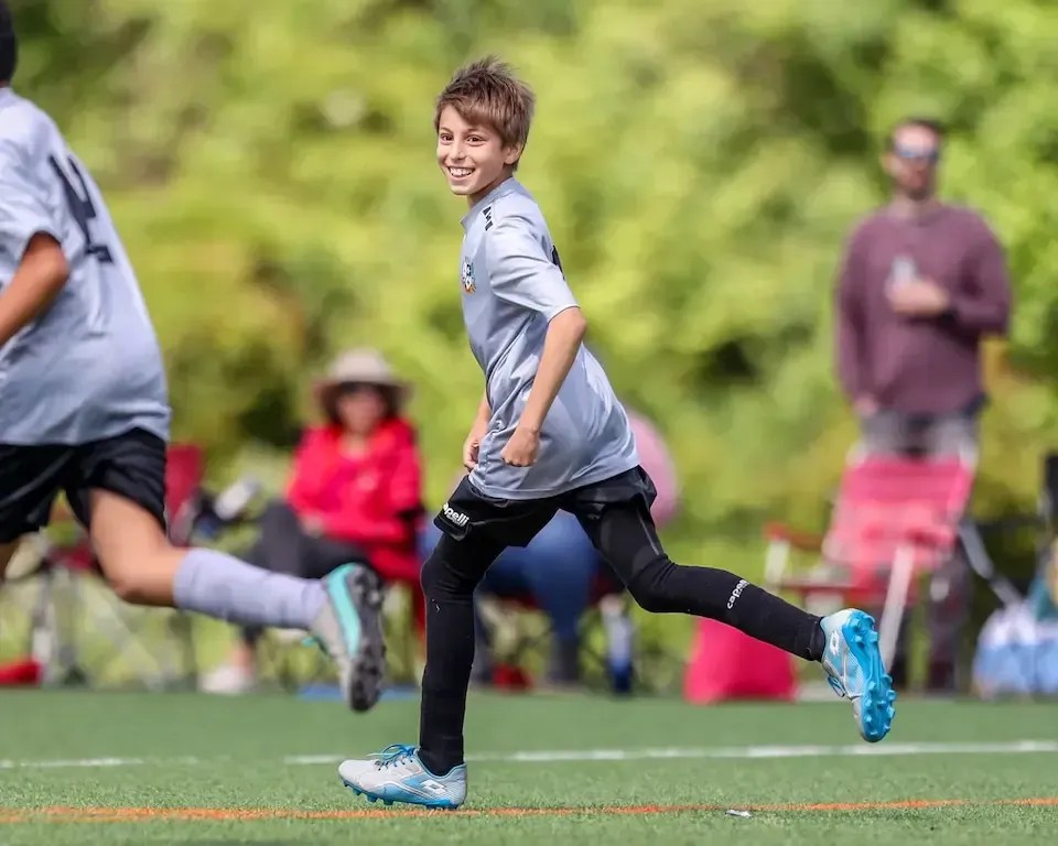 Smiling boy in gray MSI soccer uniform runs across the field during a youth soccer game with teammates and parents watching.